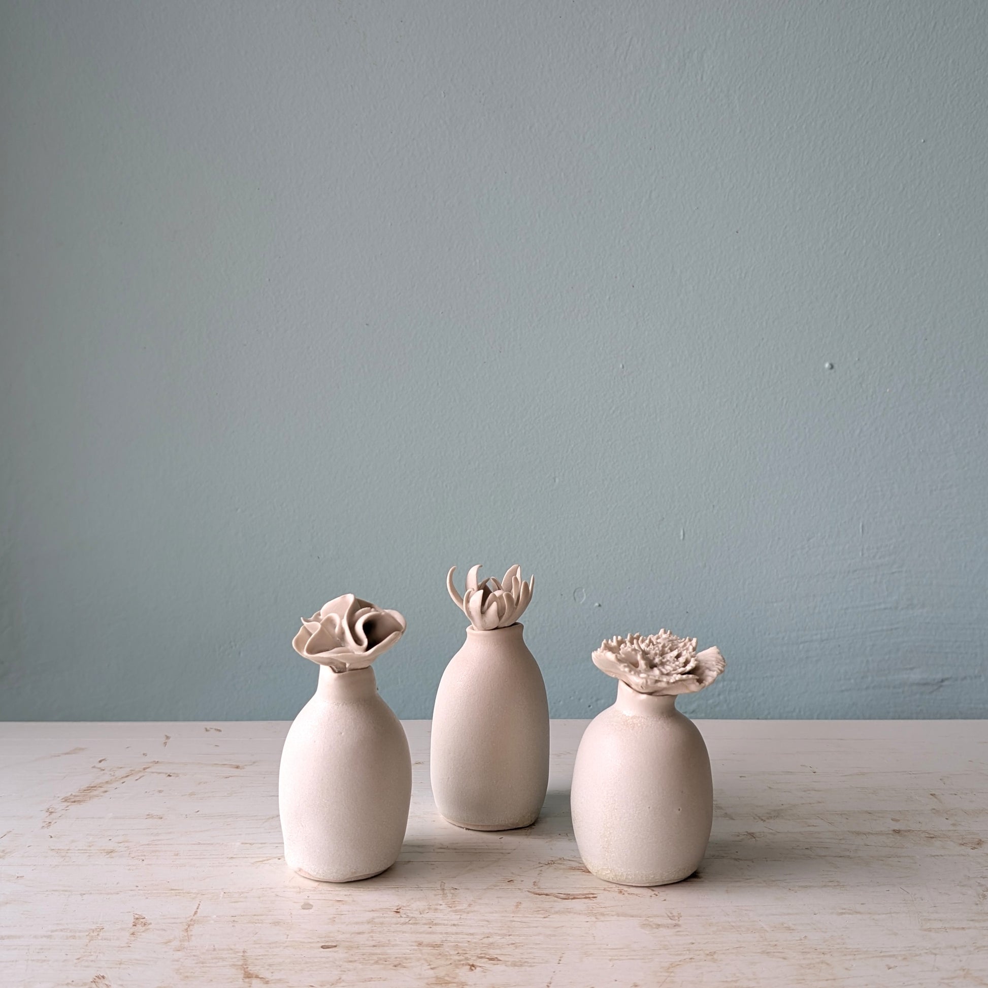 Three small matte ceramic vases in white, arranged on a light wooden surface against a pale blue wall.
