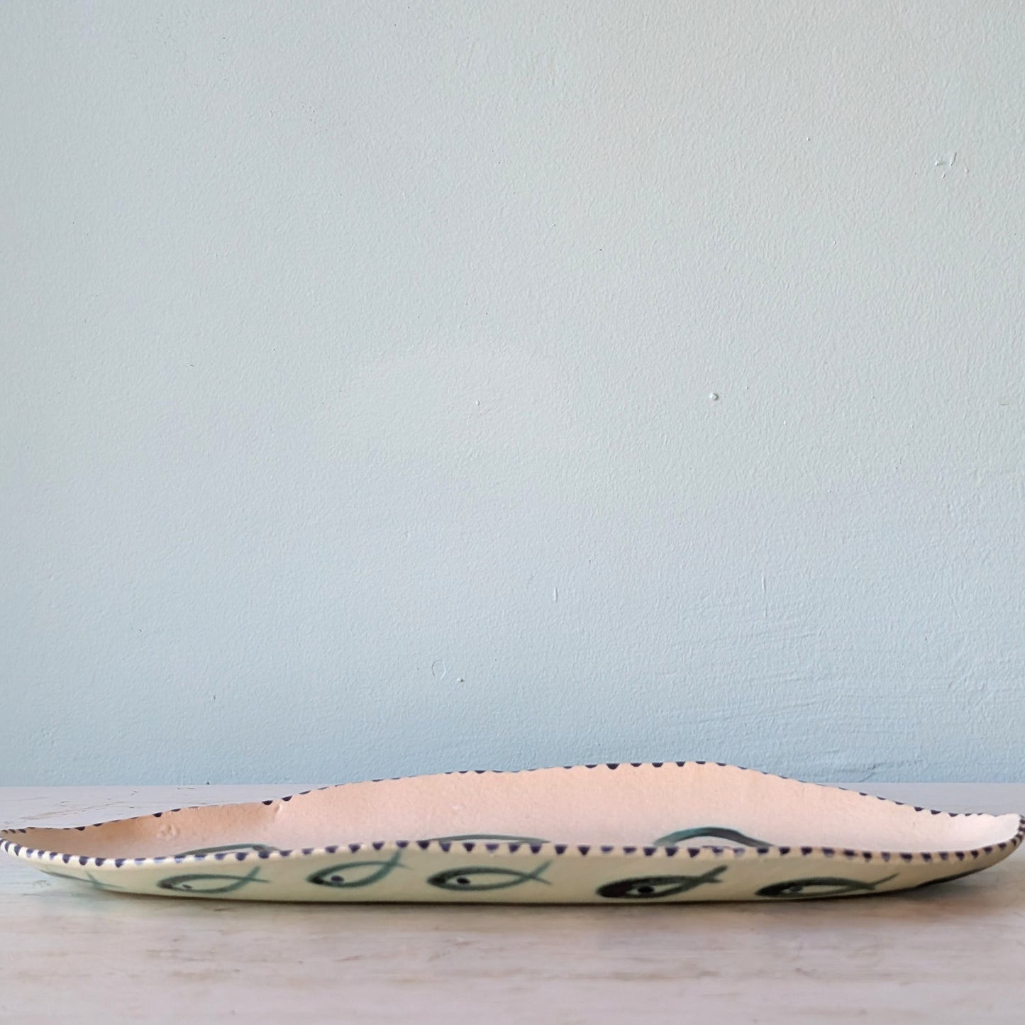 A long, shallow ceramic serving platter with a cream-colored interior and a dark green fish pattern on the exterior, resting on a light wooden surface against a pale blue wall.