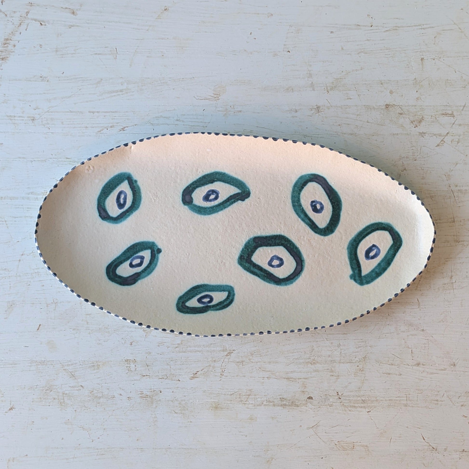 An oval ceramic platter featuring a hand-painted pattern of six dark teal teardrop shapes with white centers on a cream-colored glaze, set against a distressed white wooden background.