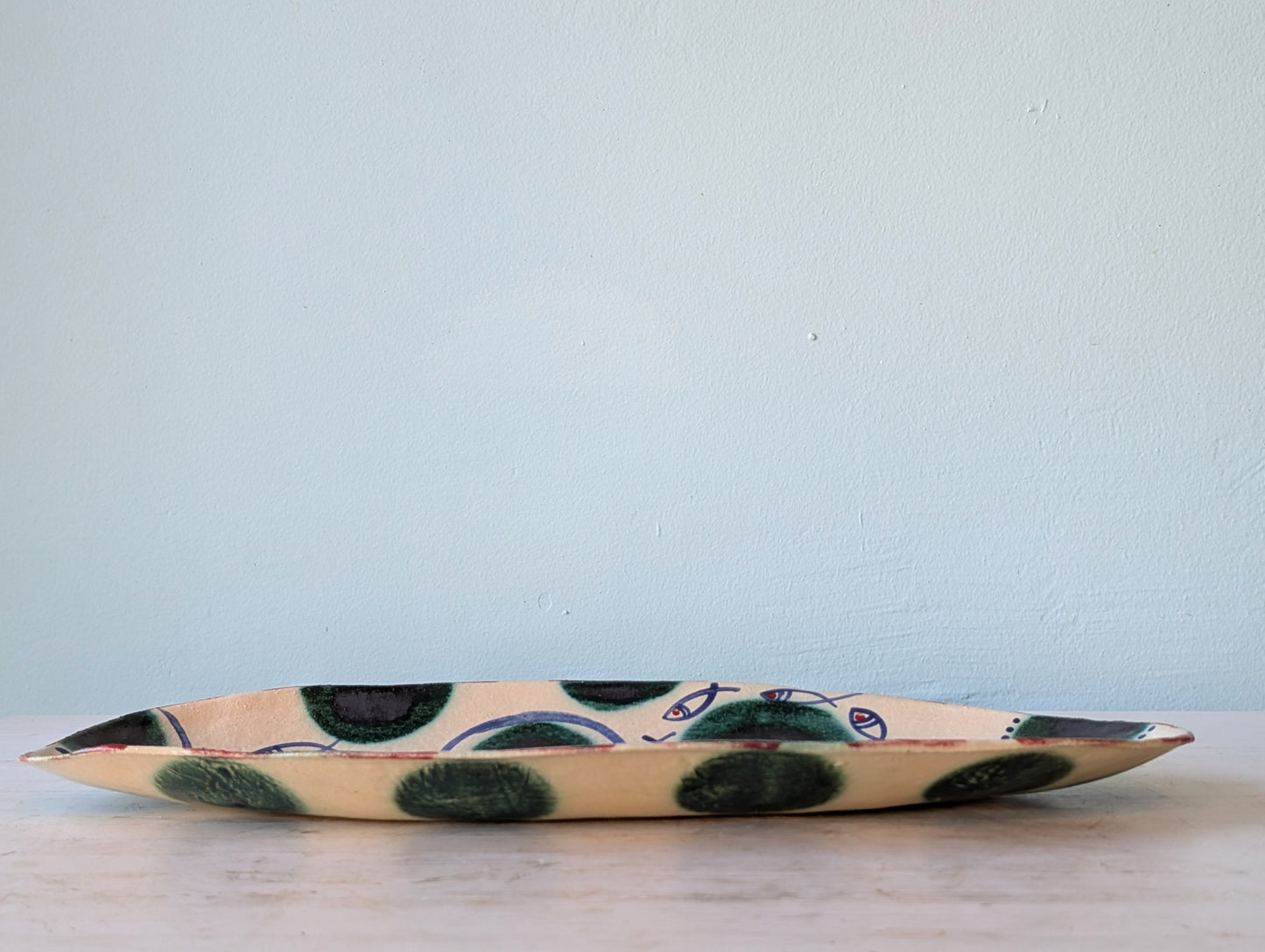 A handmade ceramic serving platter with a cream base, decorated with dark green and blue hand-painted abstract spots, resting on a light wooden surface against a pale blue wall.