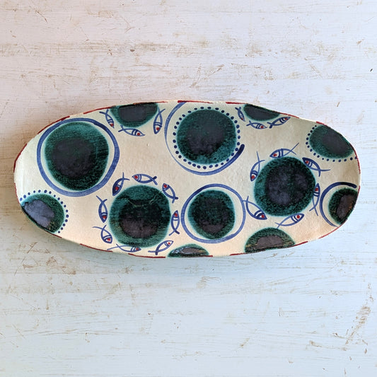 An oval ceramic serving platter with a hand-painted pattern of dark green and navy blue circles and small blue fish on a cream background, set on a distressed white wooden surface.