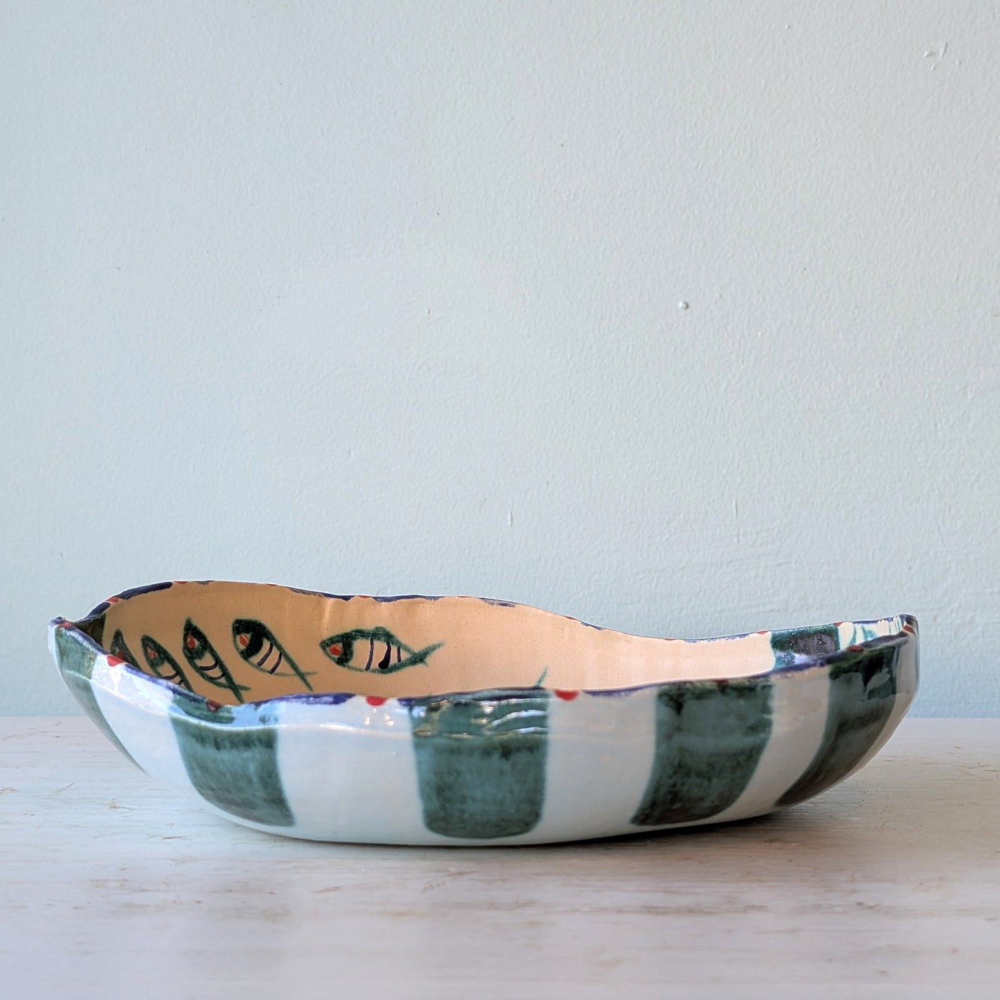 A handmade ceramic bowl with a rustic, irregular rim, featuring dark green and white vertical stripes on the exterior and painted fish designs on the interior, set against a plain light blue wall.