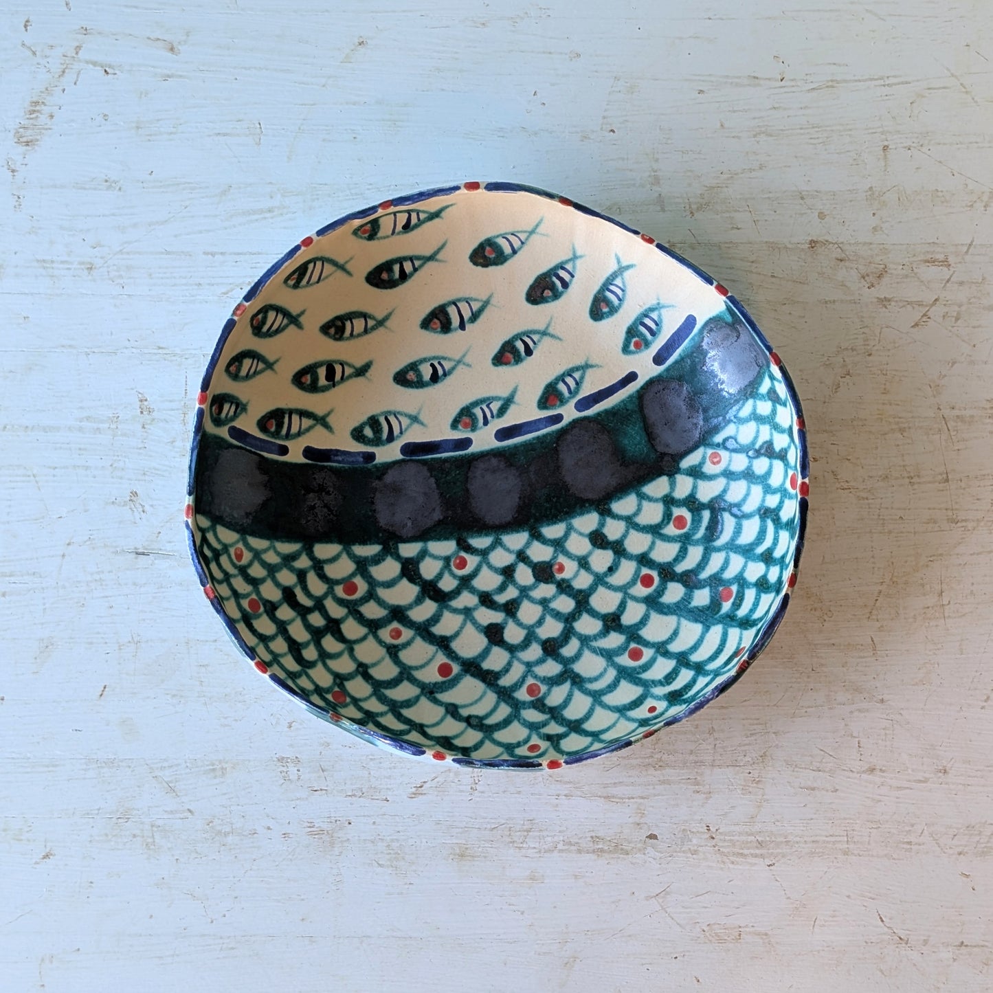 A handmade ceramic bowl with a fish pattern, photographed from above on a distressed white wooden surface.