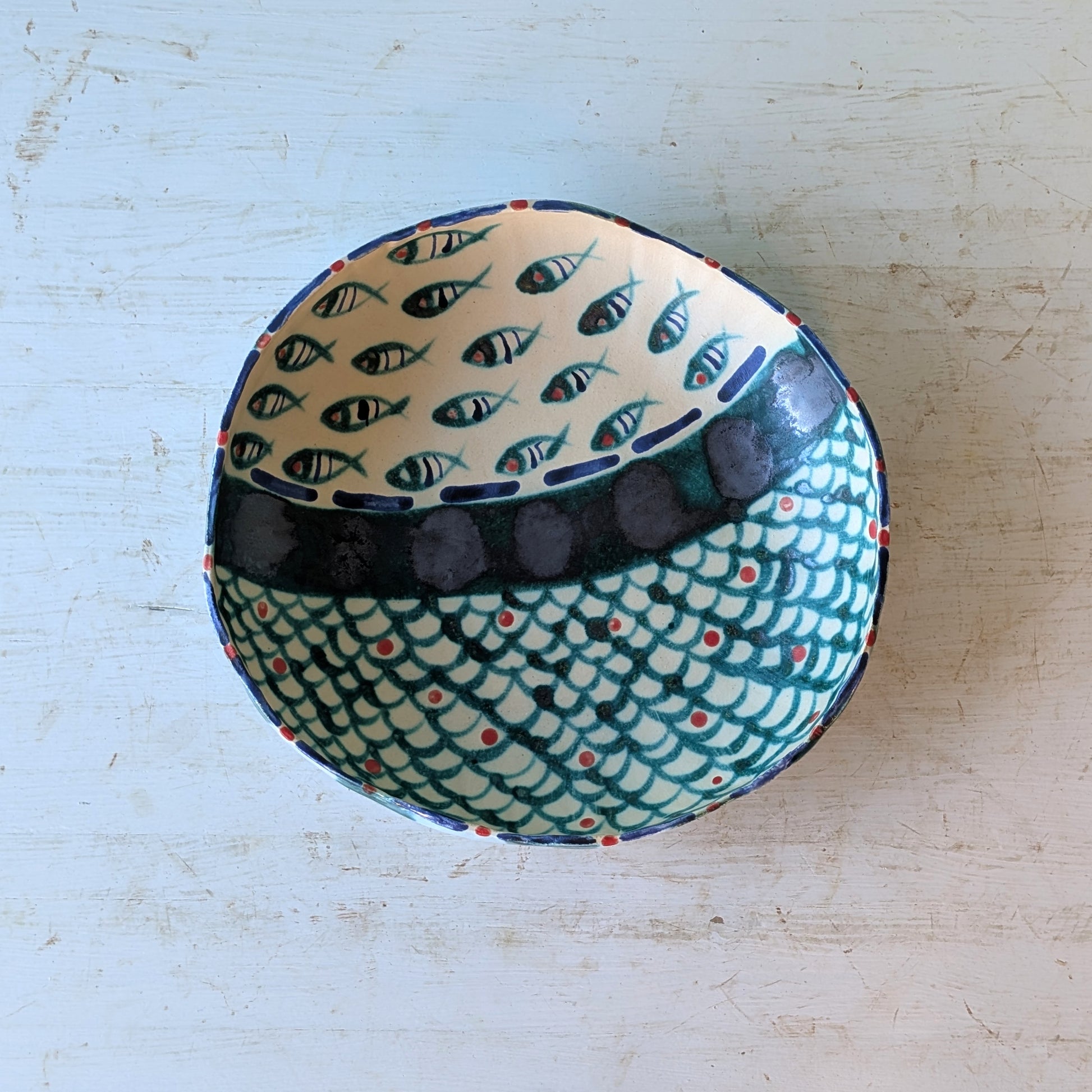 A handmade ceramic bowl with a fish pattern, photographed from above on a distressed white wooden surface.