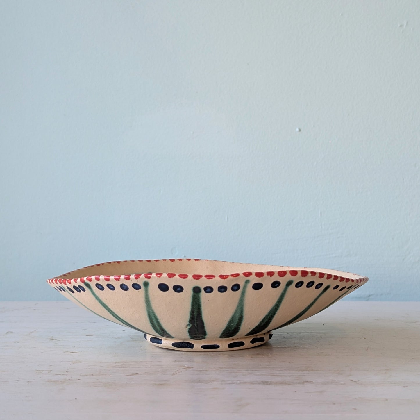 A hand-painted ceramic bowl with a cream-colored glaze, featuring green leaf-like stripes, dark green polka dots, and a red dotted rim, set against a light blue wall and white surface.