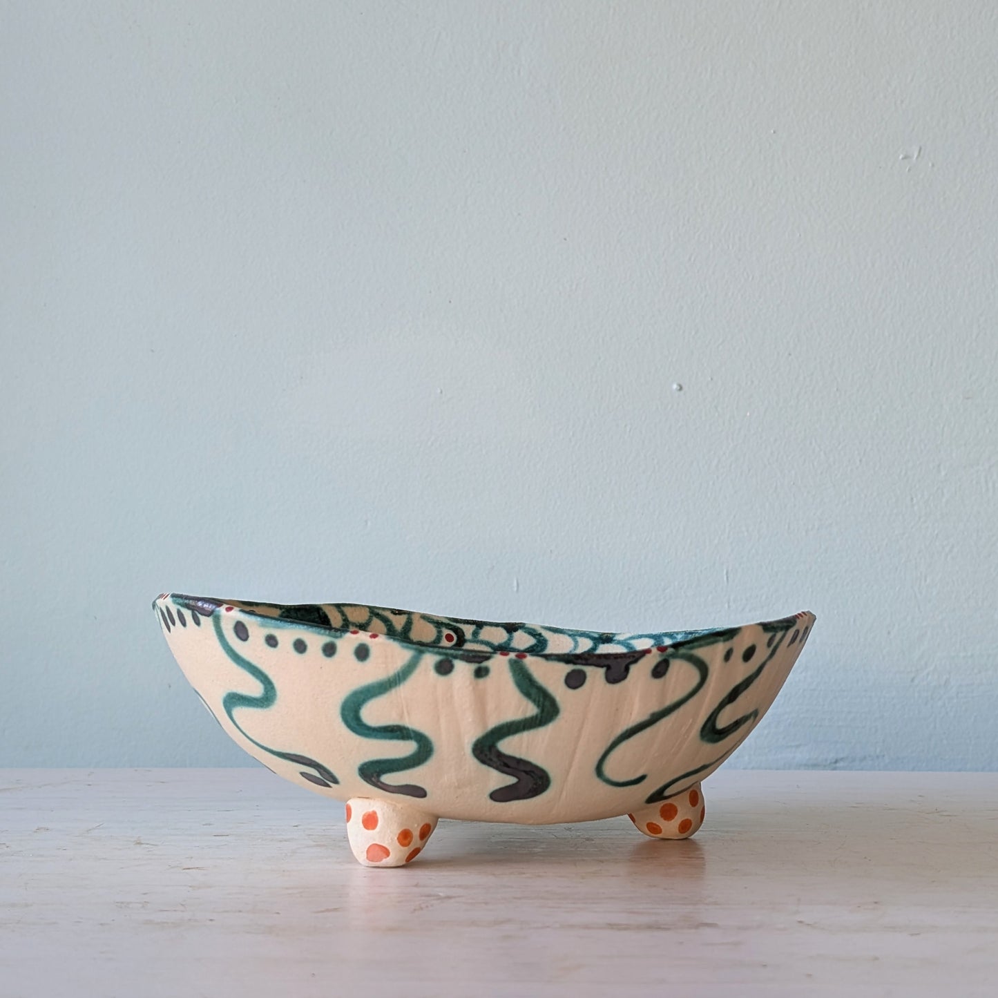 A handmade ceramic bowl with a cream-colored glaze, decorated with dark green squiggle patterns and orange polka-dot feet, set against a light blue wall.