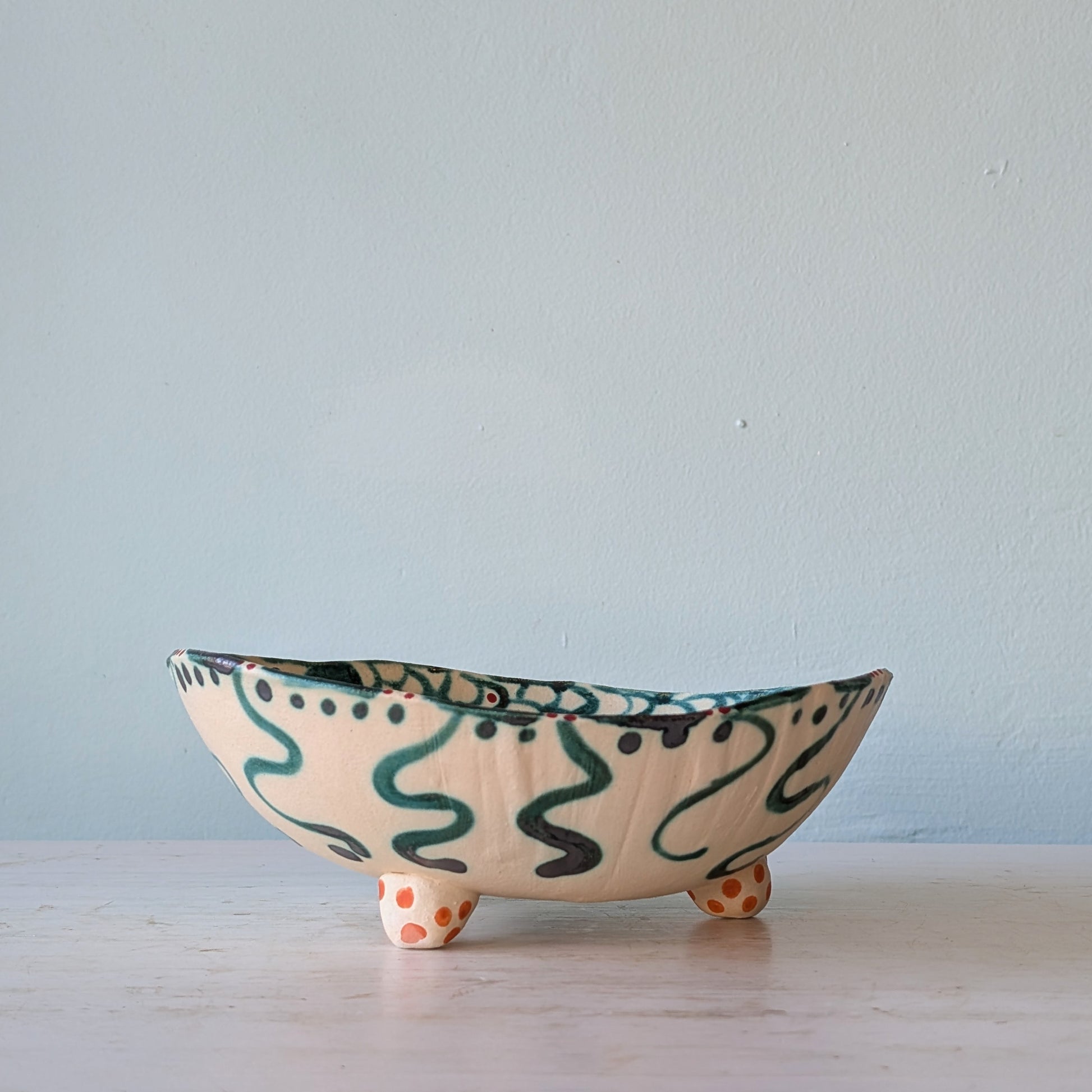A handmade ceramic bowl with a cream-colored glaze, decorated with dark green squiggle patterns and orange polka-dot feet, set against a light blue wall.
