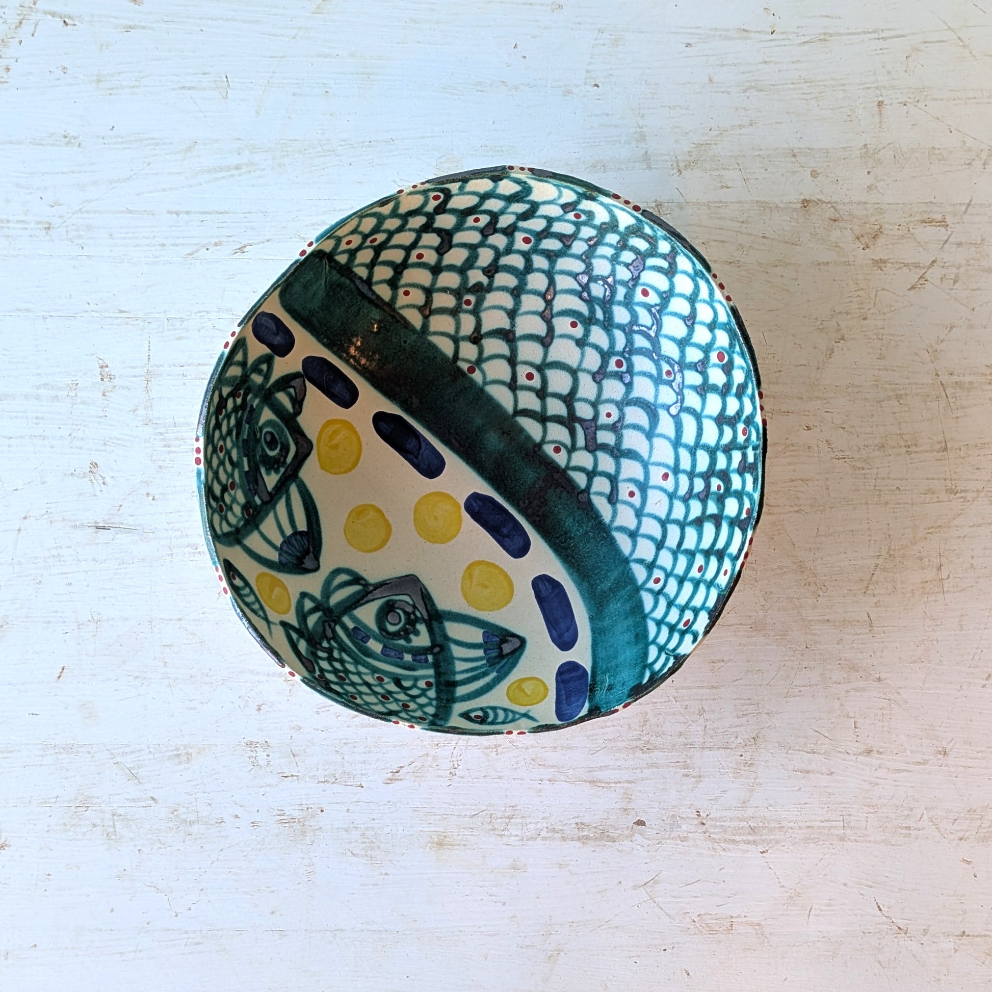 A hand-painted ceramic bowl with a white base, featuring a green fish-scale pattern, dark green fish motifs, and yellow polka dots, shown from a top-down view on a distressed white wooden surface.