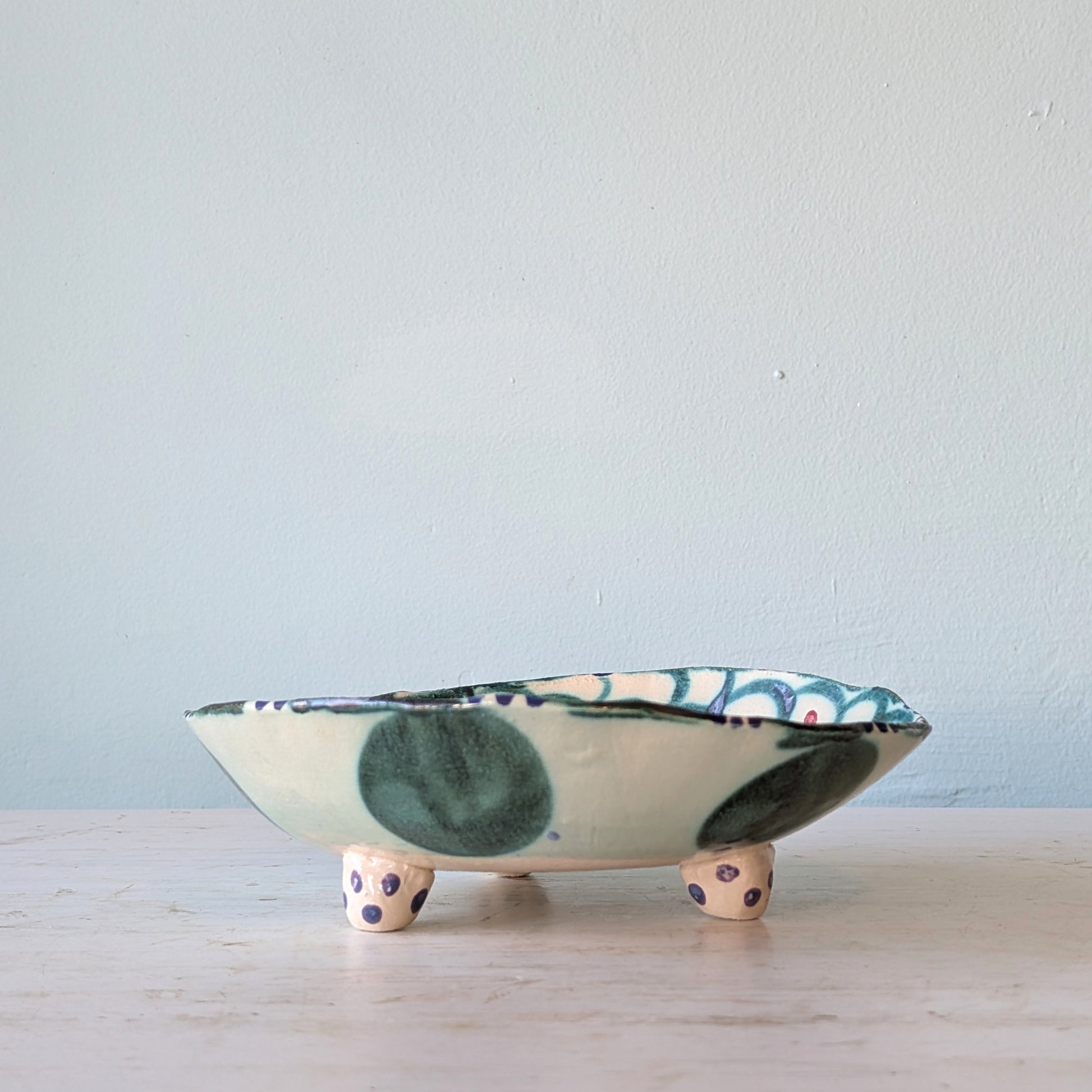 A handmade ceramic footed bowl with a cream base, dark green and blue-green painted spots, and purple polka-dot feet, set against a light blue wall.