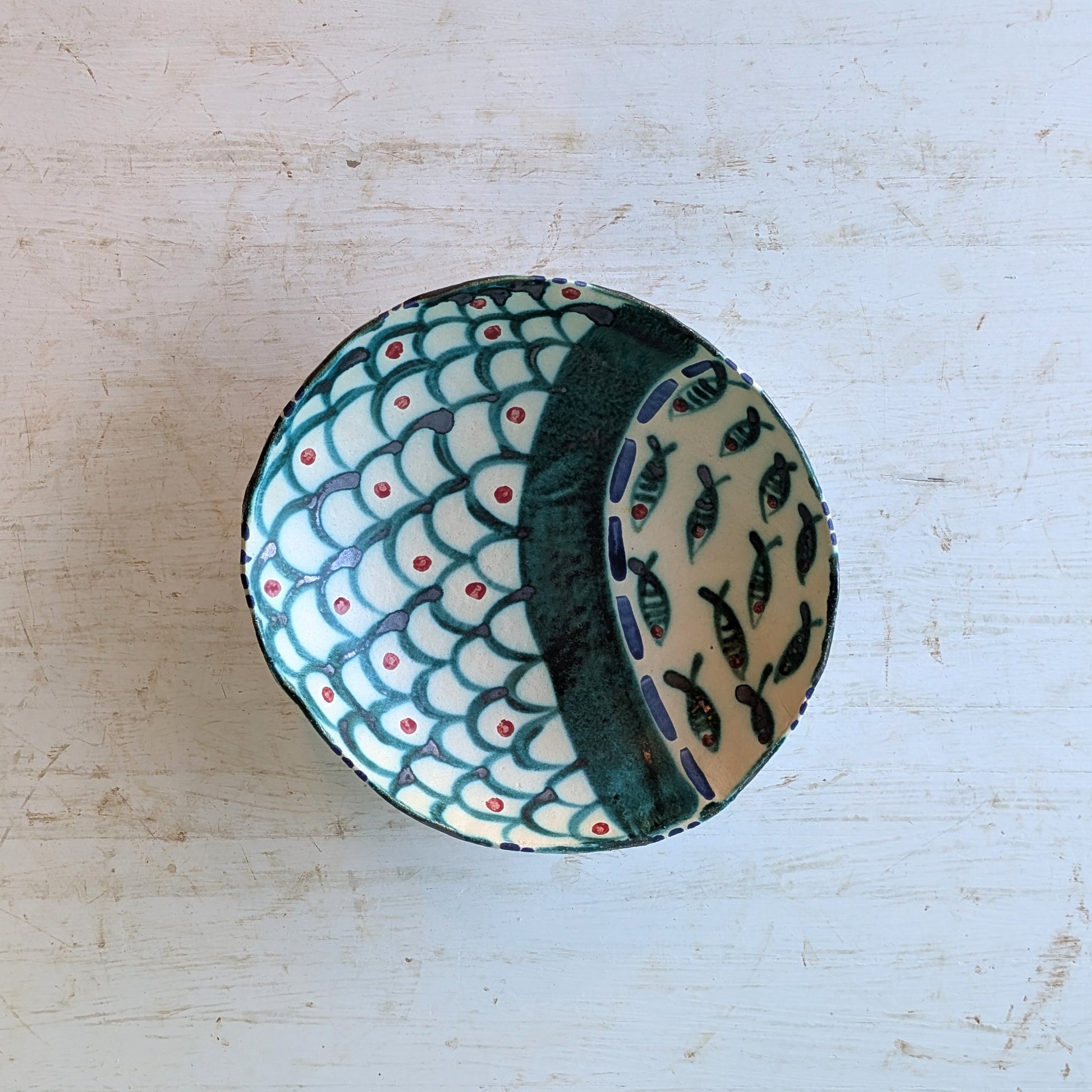 A handmade ceramic bowl with a fish-scale pattern in teal, cream, and red, and a dark green interior side with fish motifs, shown from a top-down view on a light wood background.