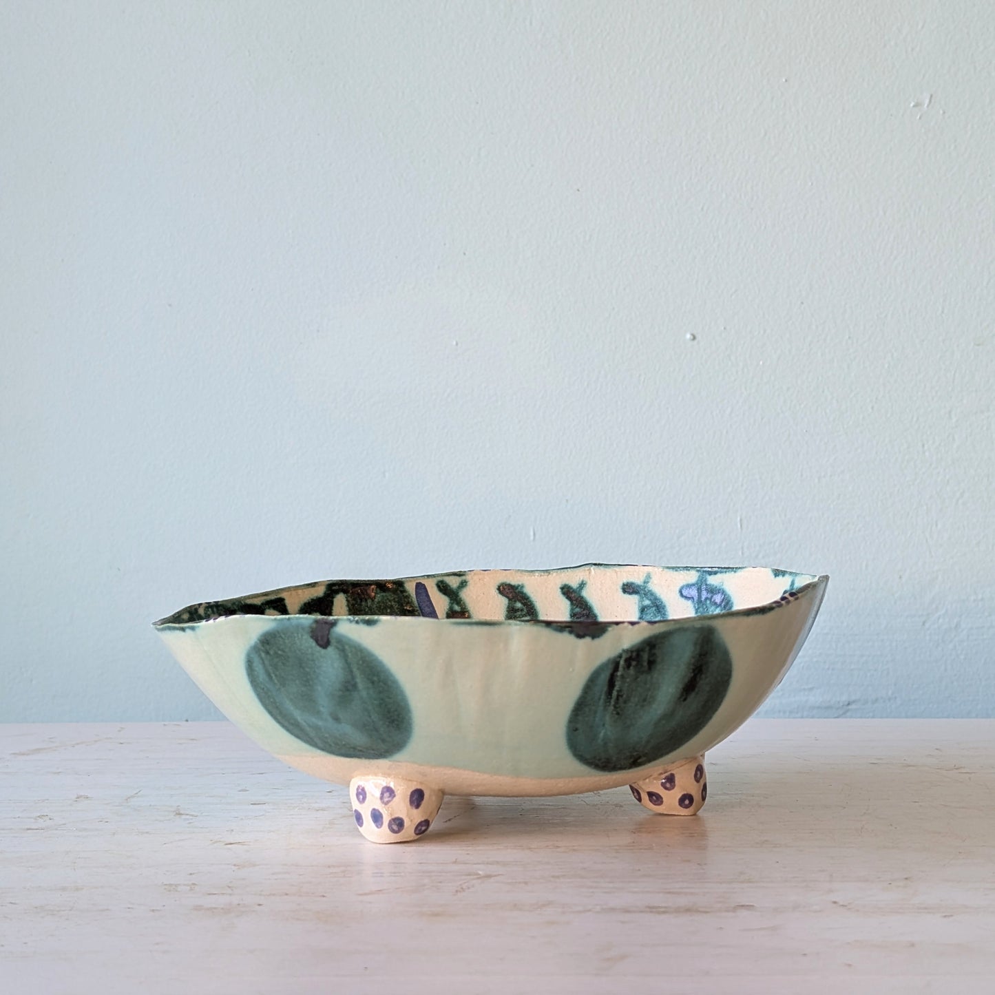 A handmade ceramic footed bowl with a light green and white glaze, decorated with dark green abstract shapes and purple polka dots on its feet, set on a light wooden surface against a pale blue wall.