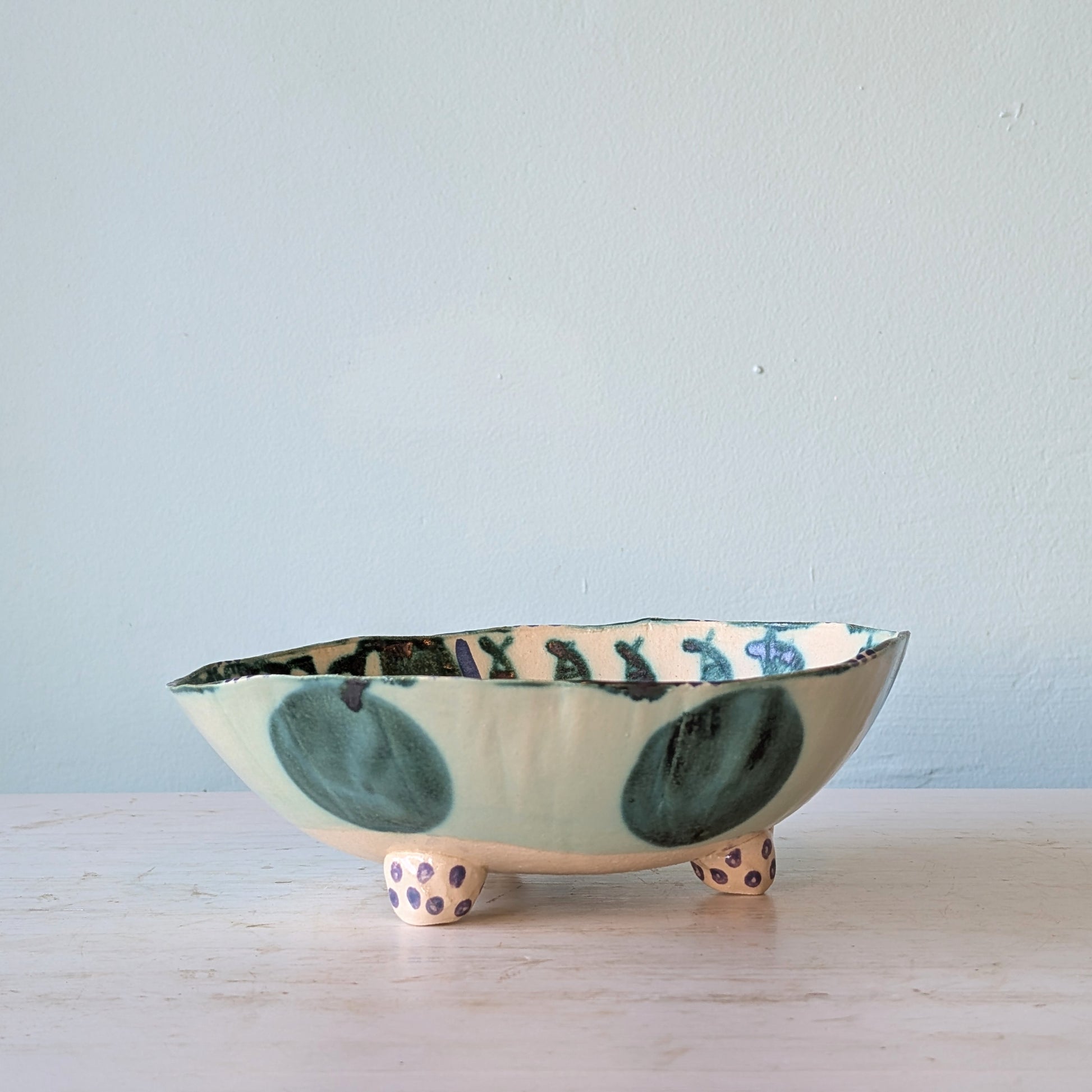 A handmade ceramic footed bowl with a light green and white glaze, decorated with dark green abstract shapes and purple polka dots on its feet, set on a light wooden surface against a pale blue wall.