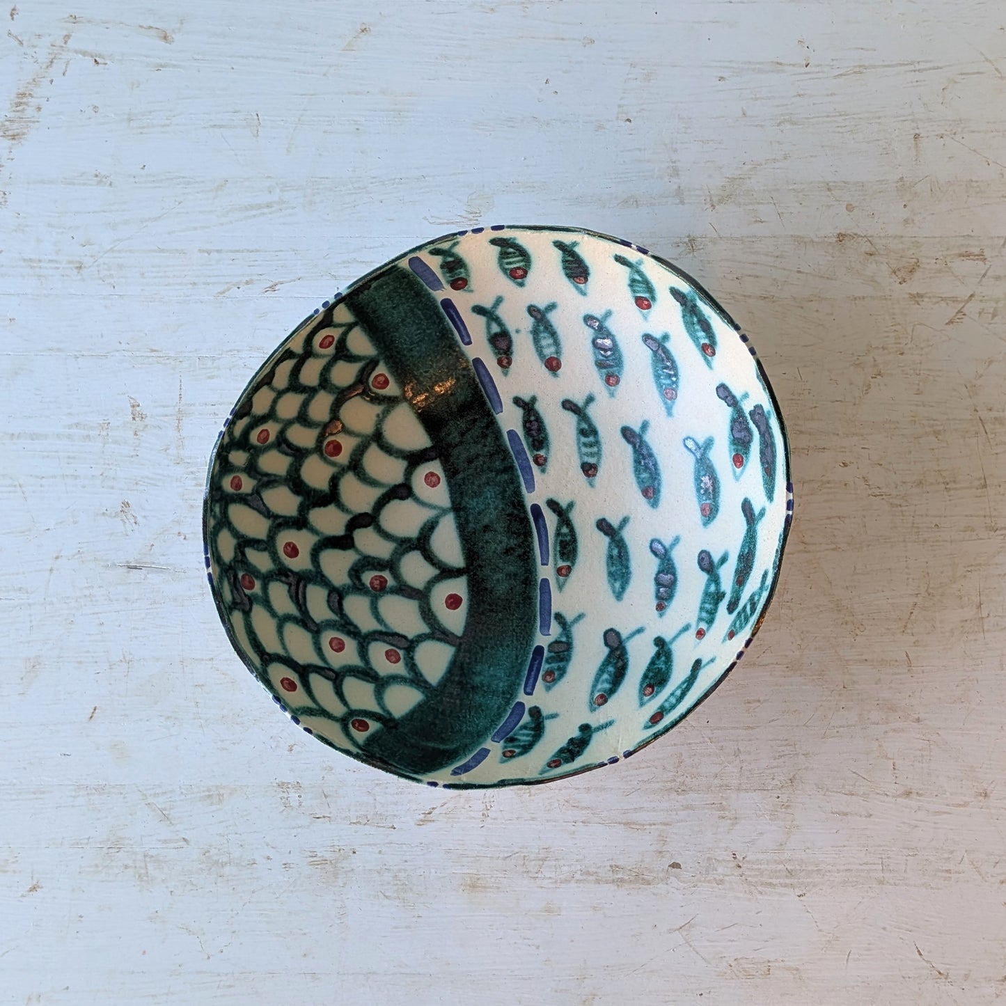 A round ceramic bowl with a white base, decorated with a pattern of small blue-green fish and a dark green scale-like design on one side, photographed from above on a distressed white wooden surface.