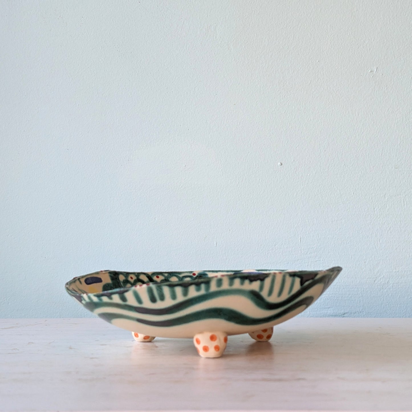 A handmade ceramic footed bowl with a cream base, dark green wavy stripes, and orange polka-dot feet, set against a light blue wall and white wooden surface.