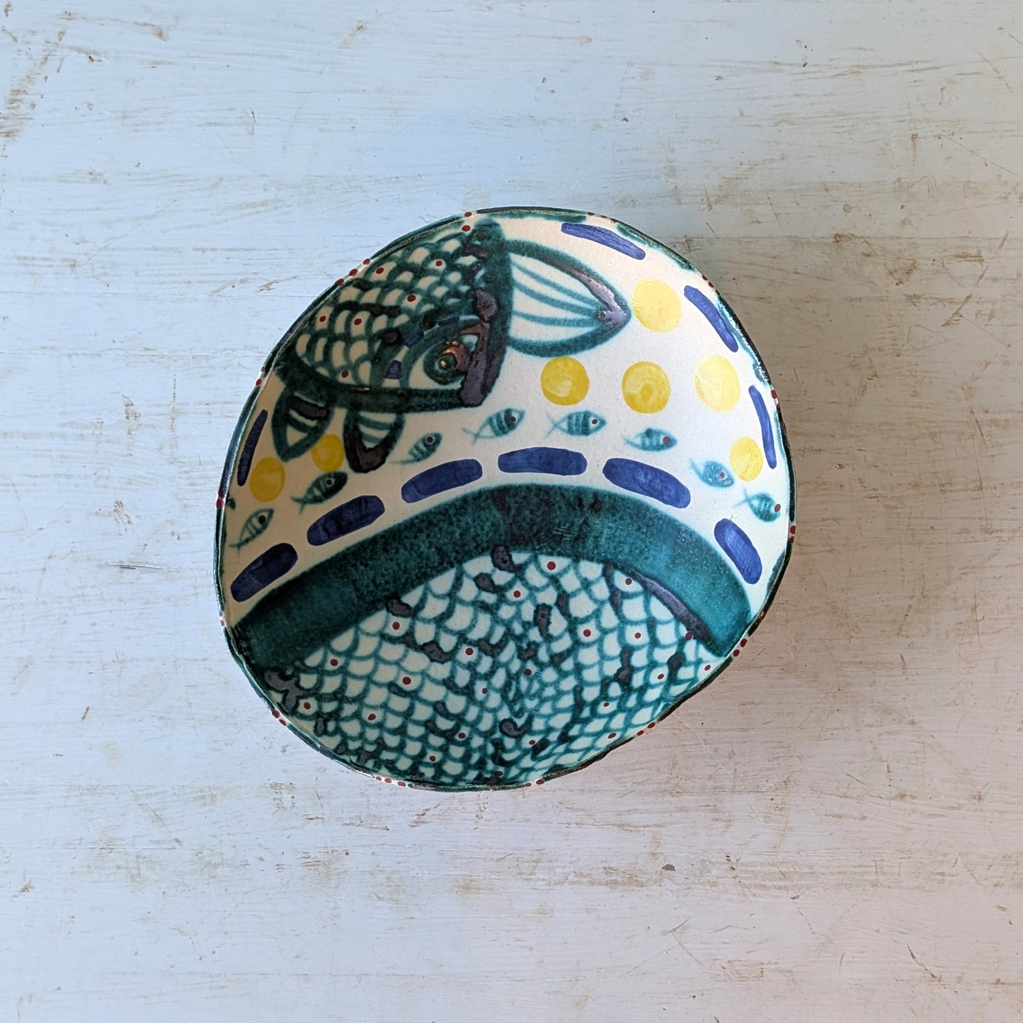 A handmade ceramic bowl with a hand-painted fish design in teal, navy, yellow, and white, shown from a top-down perspective on a distressed white wooden surface.