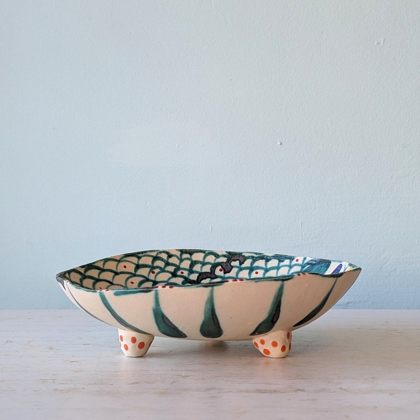 A handmade ceramic footed bowl with a cream-colored exterior, dark green painted stripes, and a green and orange fish-scale pattern on the interior, set on a light marble surface against a pale blue wall.