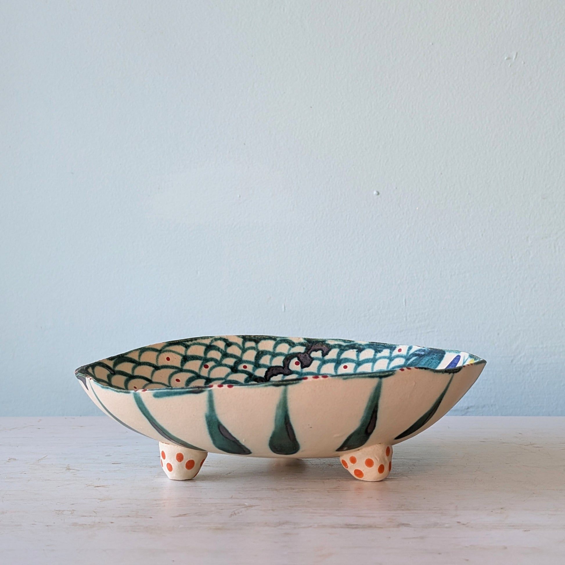 A handmade ceramic footed bowl with a cream-colored exterior, dark green painted stripes, and a green and orange fish-scale pattern on the interior, set on a light marble surface against a pale blue wall.