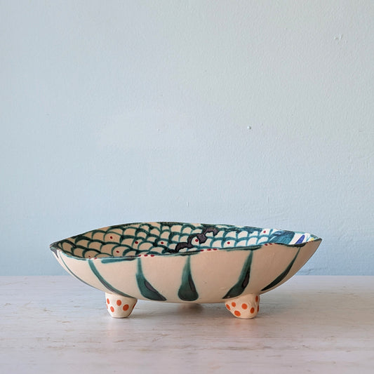 A handmade ceramic footed bowl with a cream-colored exterior, dark green painted stripes, and a green and orange fish-scale pattern on the interior, set on a light marble surface against a pale blue wall.