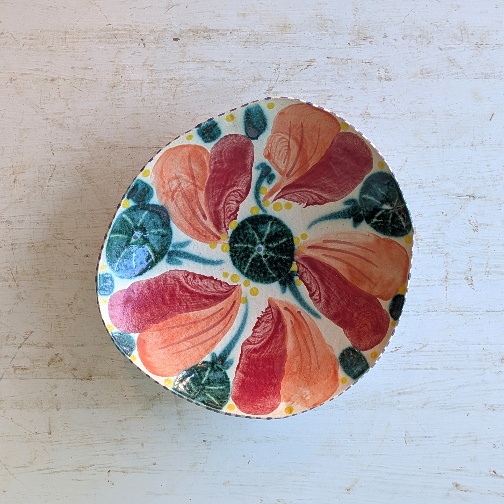 A round ceramic bowl with a hand-painted floral pattern featuring orange, red, and green petals on a cream background, set on a distressed white wooden surface.