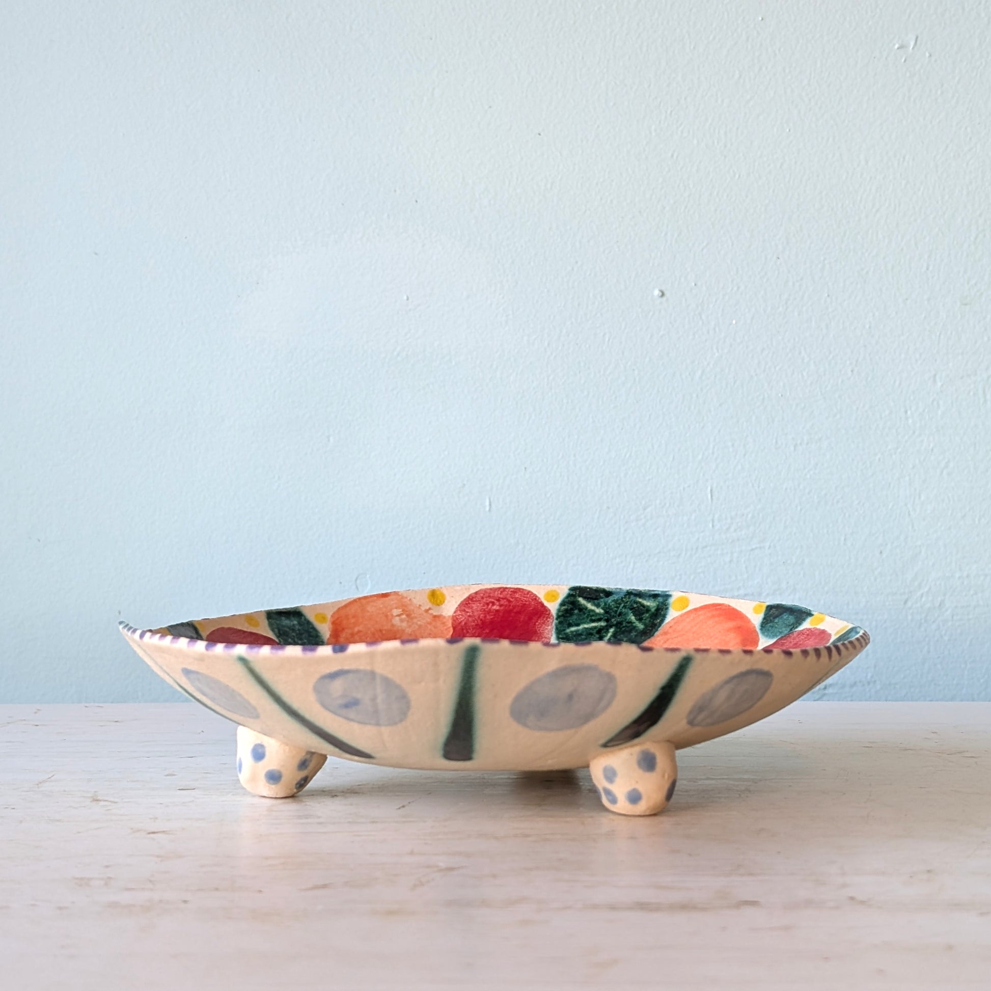 A handmade ceramic footed bowl with a colorful fruit and leaf pattern, resting on a light wooden surface against a pale blue wall.