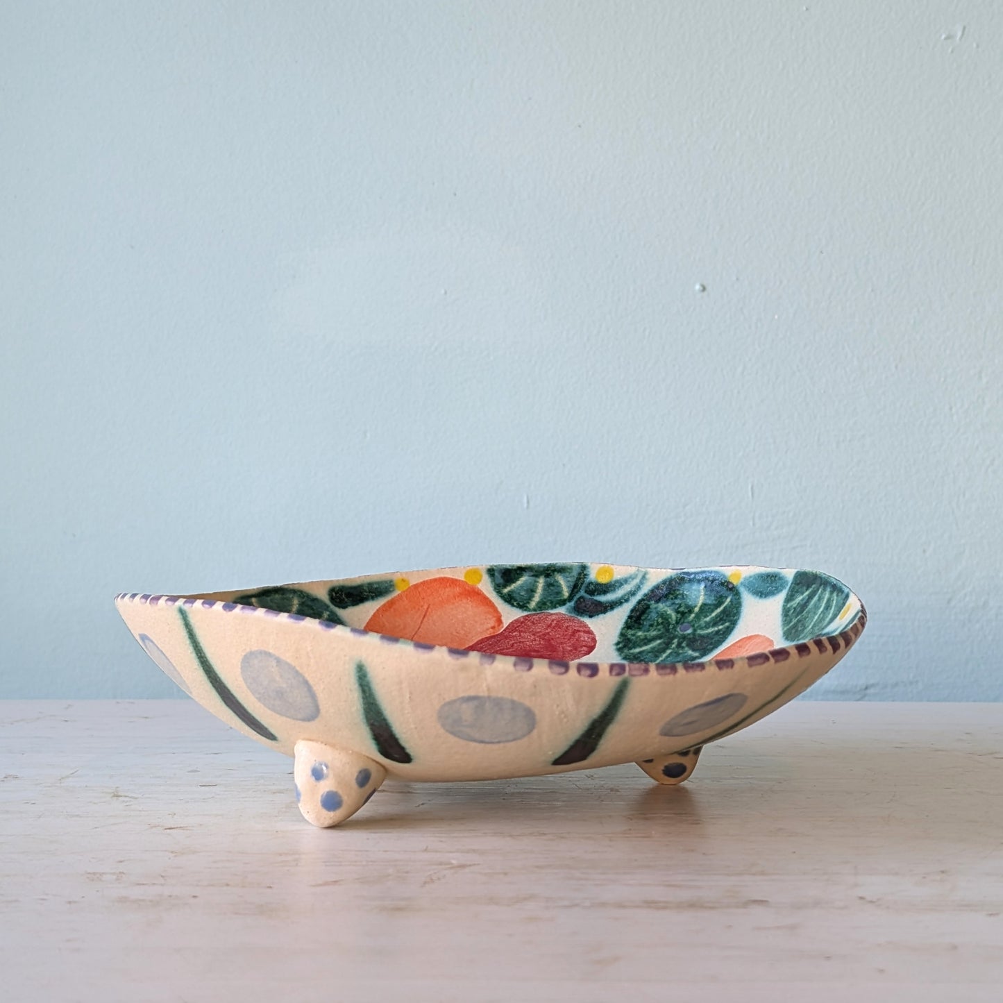 A handmade ceramic footed bowl with a cream-colored base, decorated with colorful painted motifs of green leaves, orange and red fruits, and gray polka dots, set on a light wooden surface against a pale blue wall.