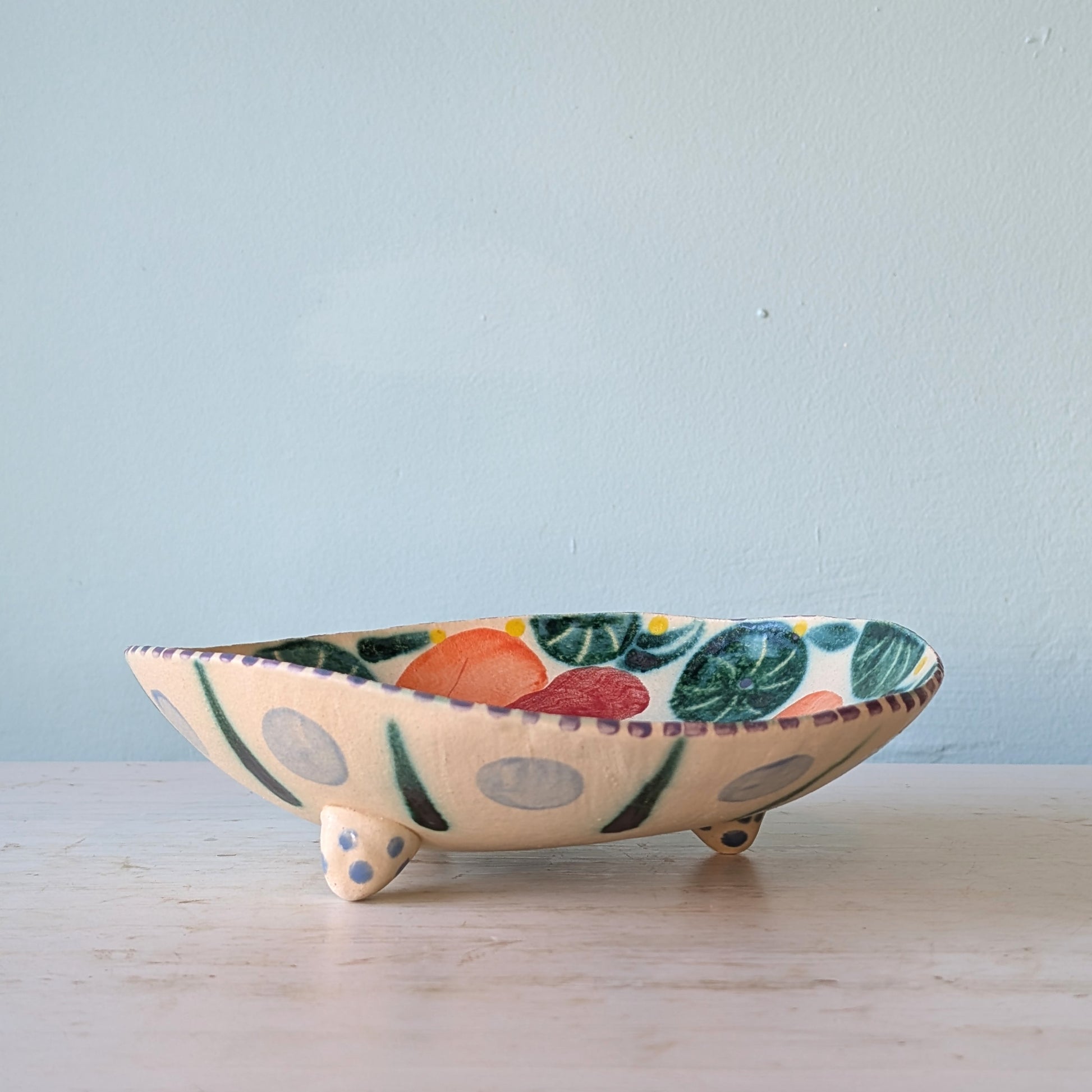A handmade ceramic footed bowl with a cream-colored base, decorated with colorful painted motifs of green leaves, orange and red fruits, and gray polka dots, set on a light wooden surface against a pale blue wall.