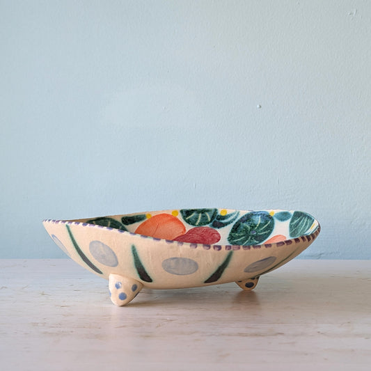 A handmade ceramic footed bowl with a cream-colored base, decorated with colorful painted motifs of green leaves, orange and red fruits, and gray polka dots, set on a light wooden surface against a pale blue wall.