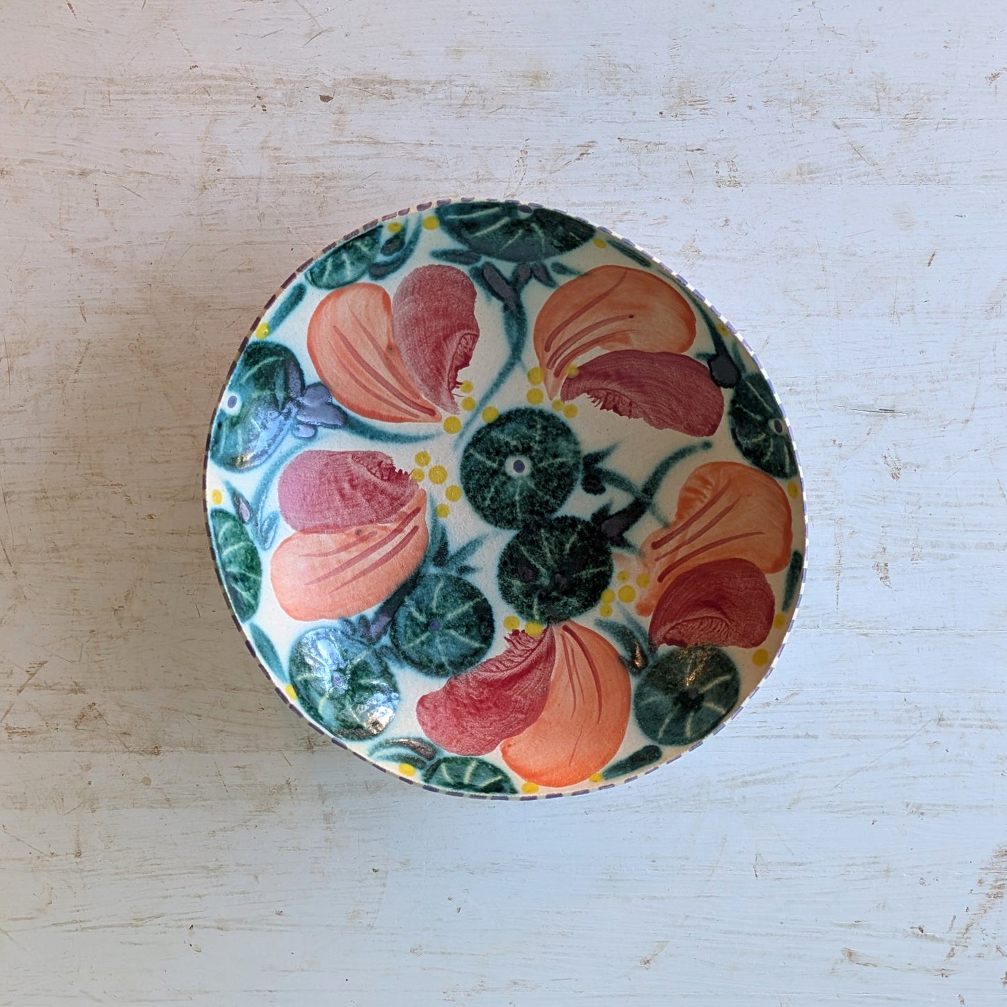 A round ceramic bowl with a colorful floral pattern featuring orange, yellow, and green leaves on a cream background, photographed from above on a distressed white wooden surface.