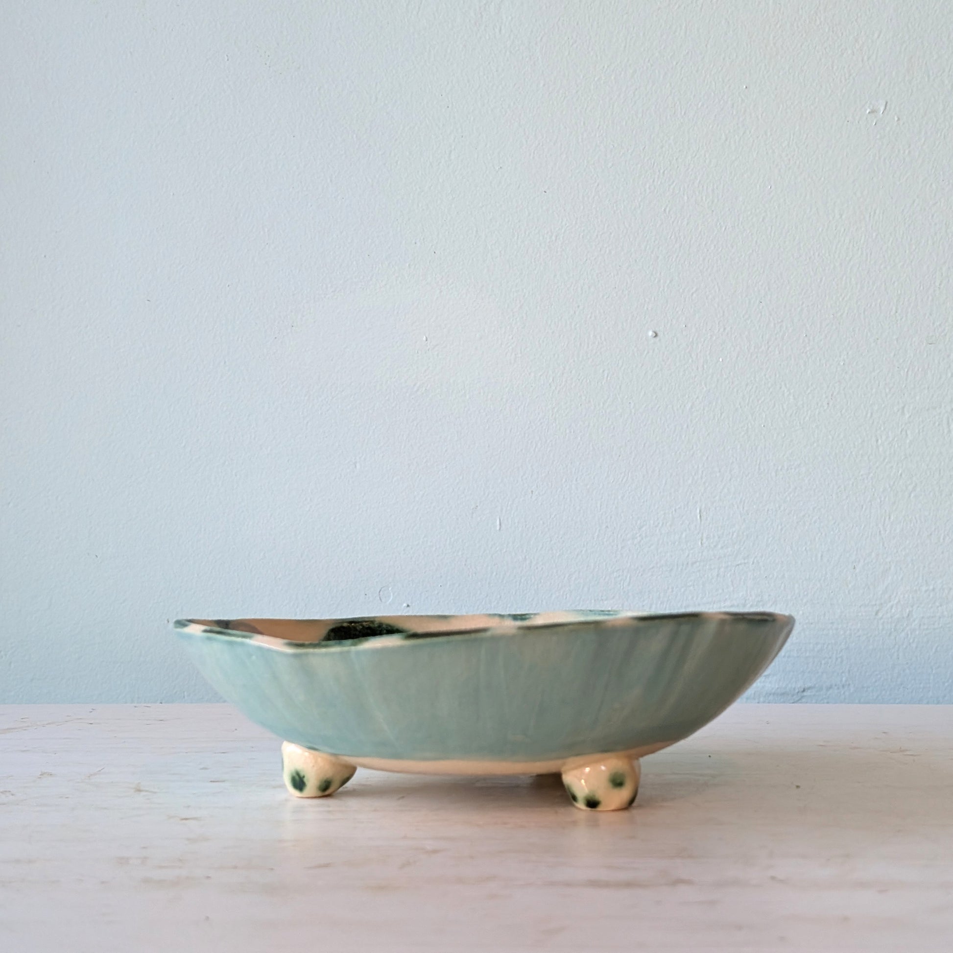 A shallow ceramic bowl with a pale sage green glaze and small cream-colored feet, set against a light blue wall and a light wooden surface.