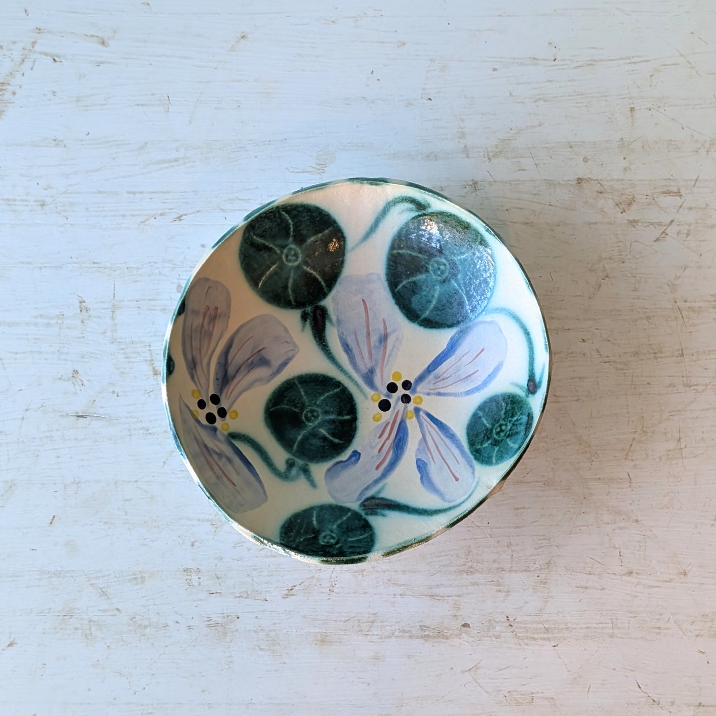 A round ceramic bowl with a hand-painted floral design featuring blue and yellow flowers with green leaves on a cream background, placed on a distressed white wooden surface.