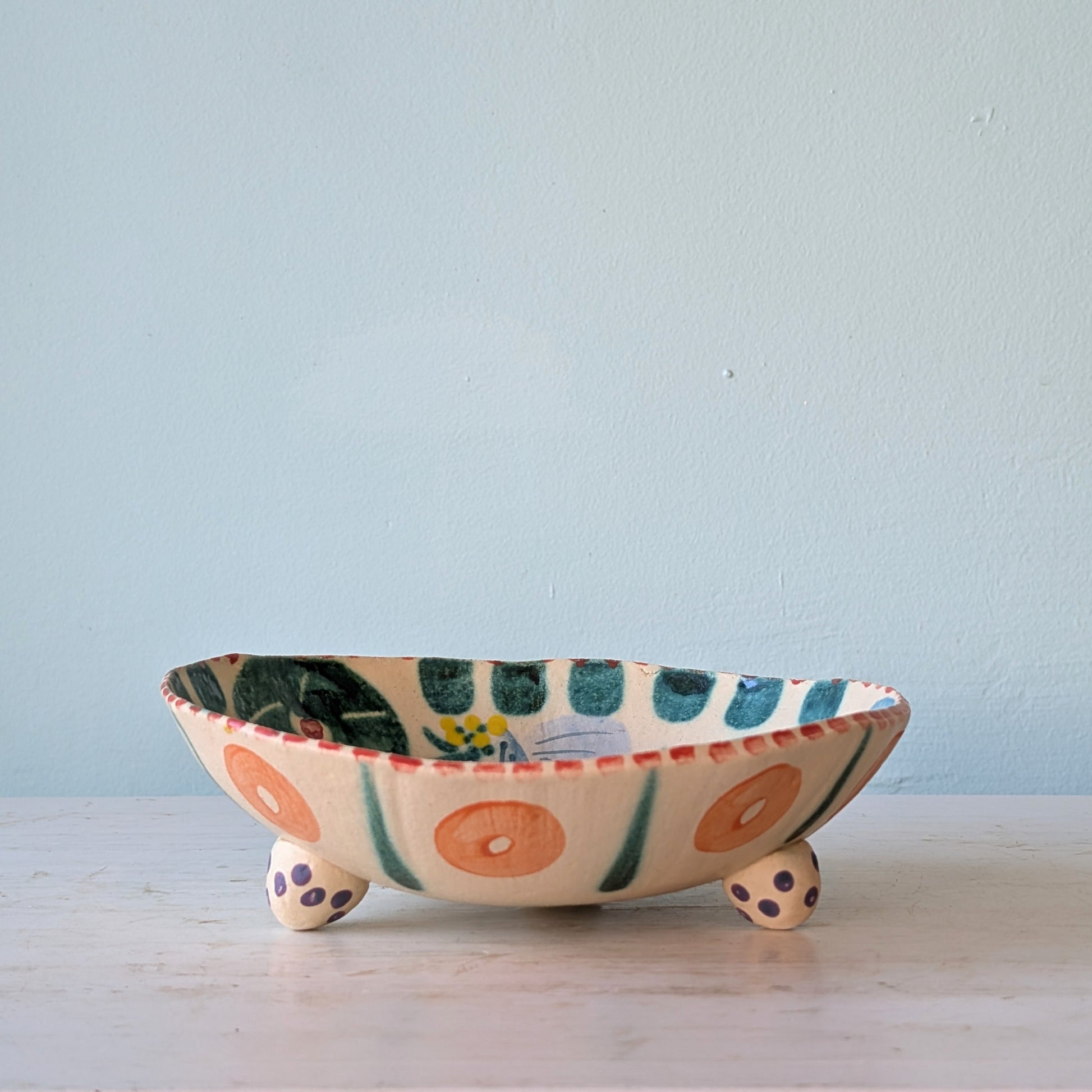 A handmade ceramic bowl with three small legs, decorated with hand-painted orange circles, green stripes, and blue dots, sitting on a light-colored wooden surface against a pale blue wall.