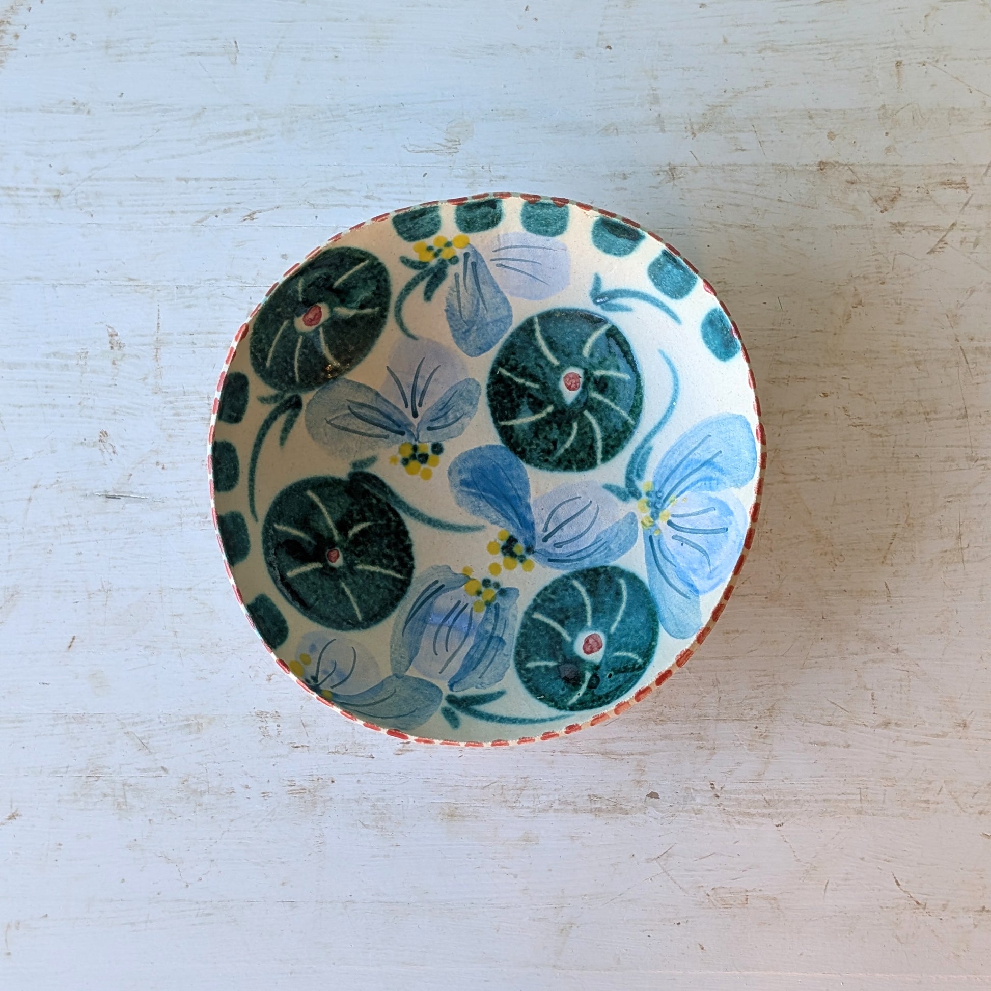 A round ceramic bowl with a hand-painted floral pattern featuring blue and green flowers on a white background, set on a distressed white wooden surface.