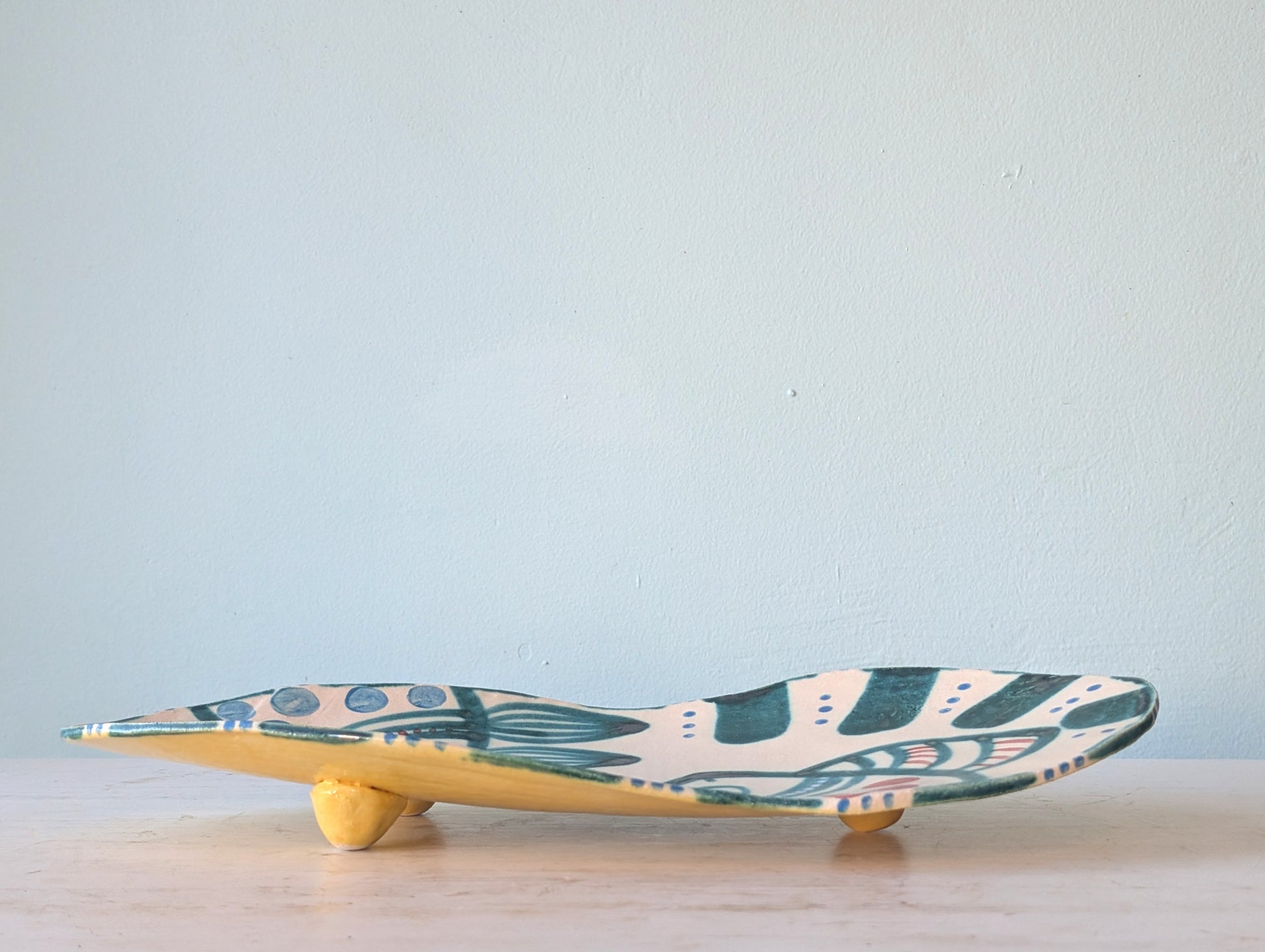 A handcrafted ceramic platter with a wavy, elongated shape, featuring a cream-colored top decorated with teal and pink abstract patterns, and a yellow glazed underside, resting on a light wooden surface against a pale blue wall.