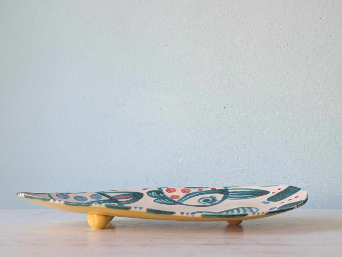 A handmade ceramic tray with a whimsical hand-painted design, featuring a cream background with green, blue, and orange patterns, supported by two small yellow feet on a light wooden surface against a pale blue wall.