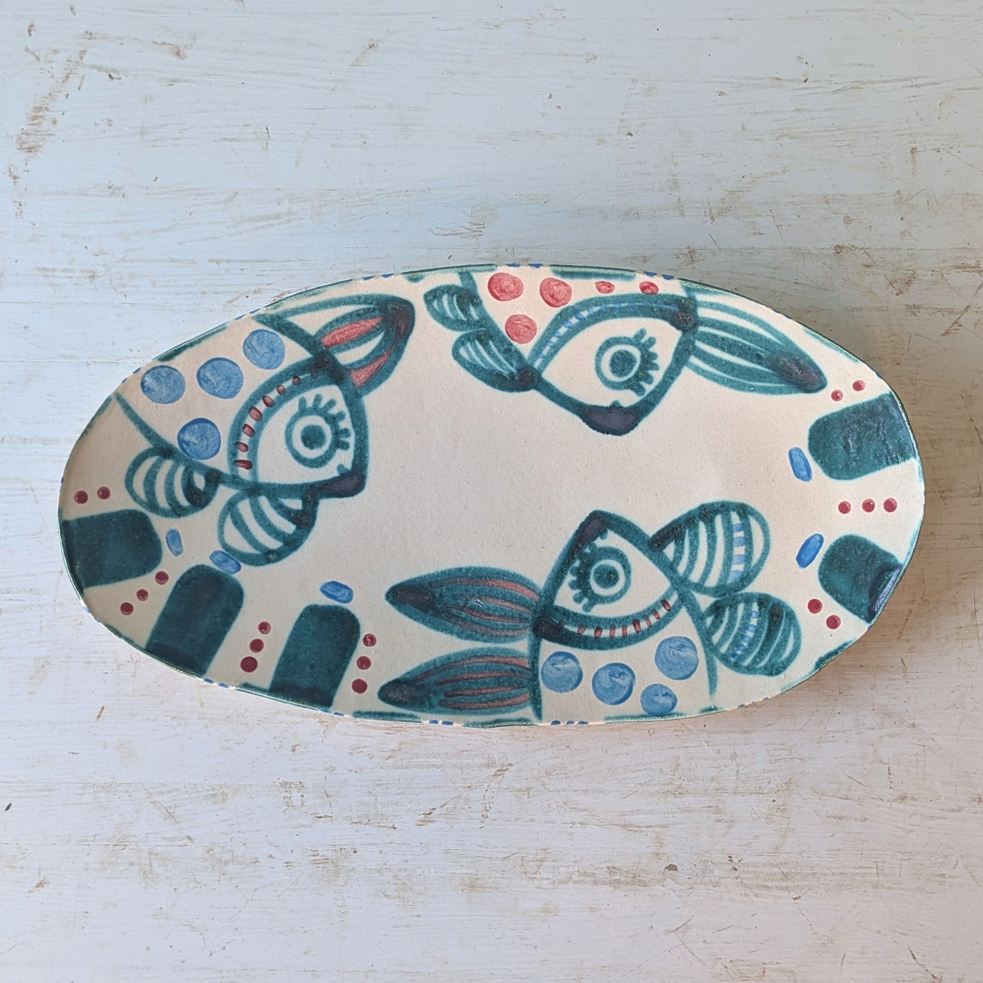 An oval ceramic serving platter with a hand-painted folk art design of stylized fish in teal, blue, and red on an off-white background, set on a distressed white wooden surface.