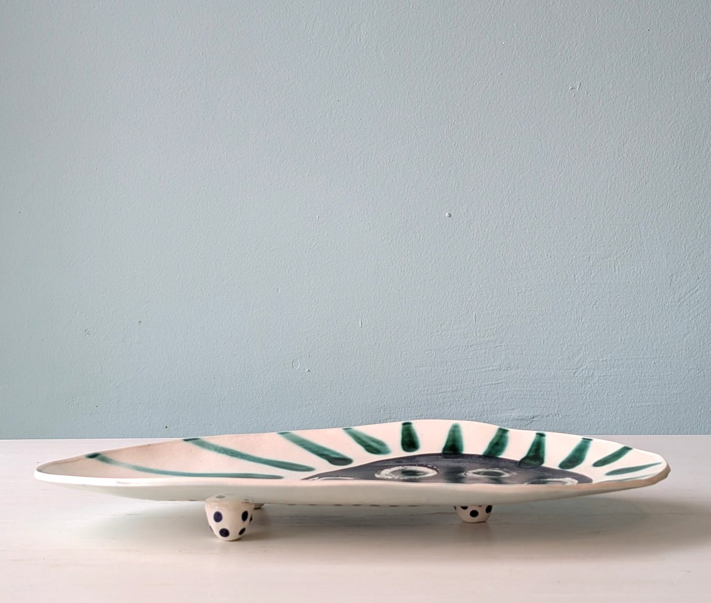 Front view of a footed white ceramic oval platter with green stripes on a white table against a blue wall