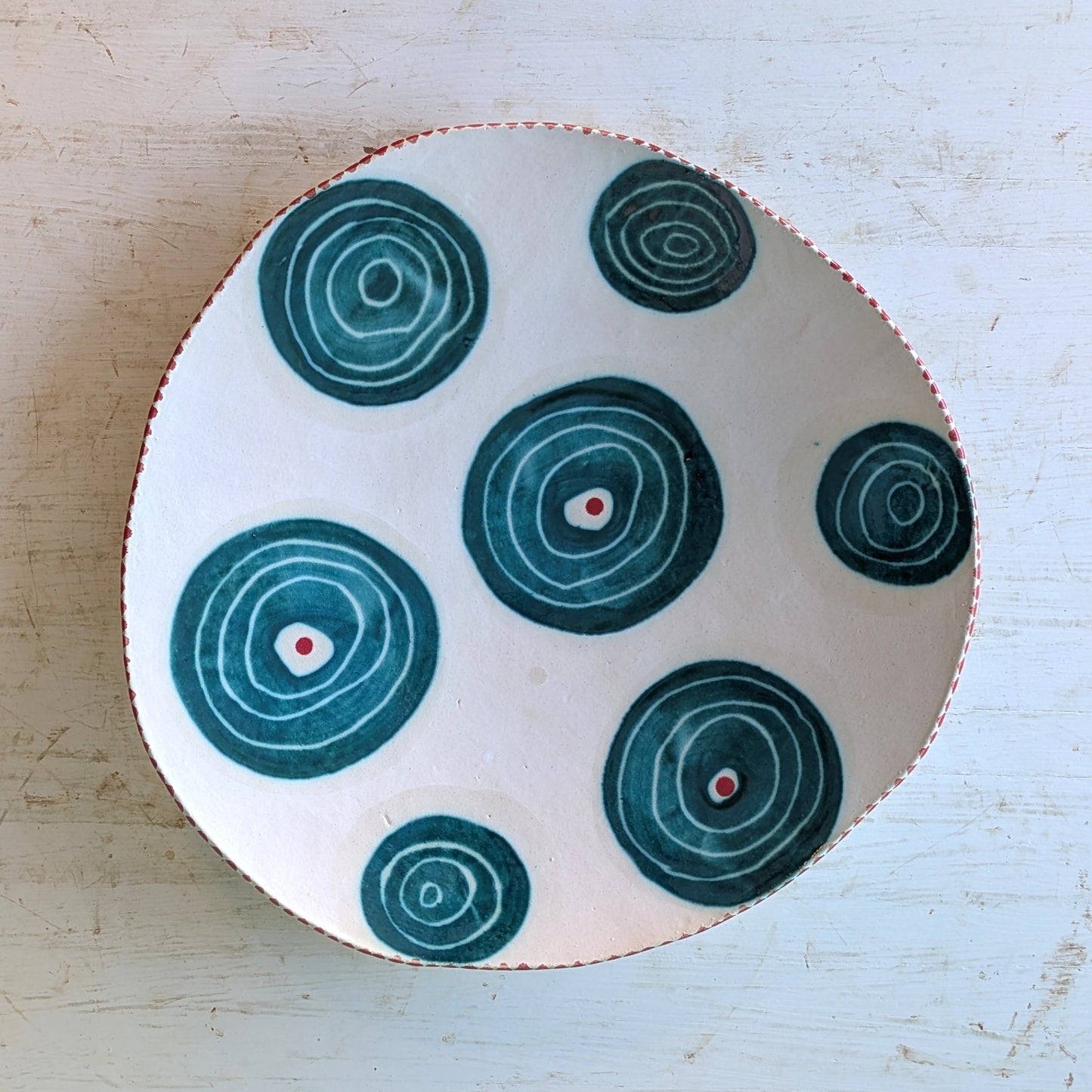 A handmade ceramic platter with a hand-drawn organic shape, featuring a white base decorated with dark teal concentric circle patterns and small red dots, set on a light weathered wood background.