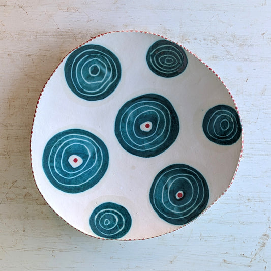 A handmade ceramic platter with a hand-drawn organic shape, featuring a white base decorated with dark teal concentric circle patterns and small red dots, set on a light weathered wood background.