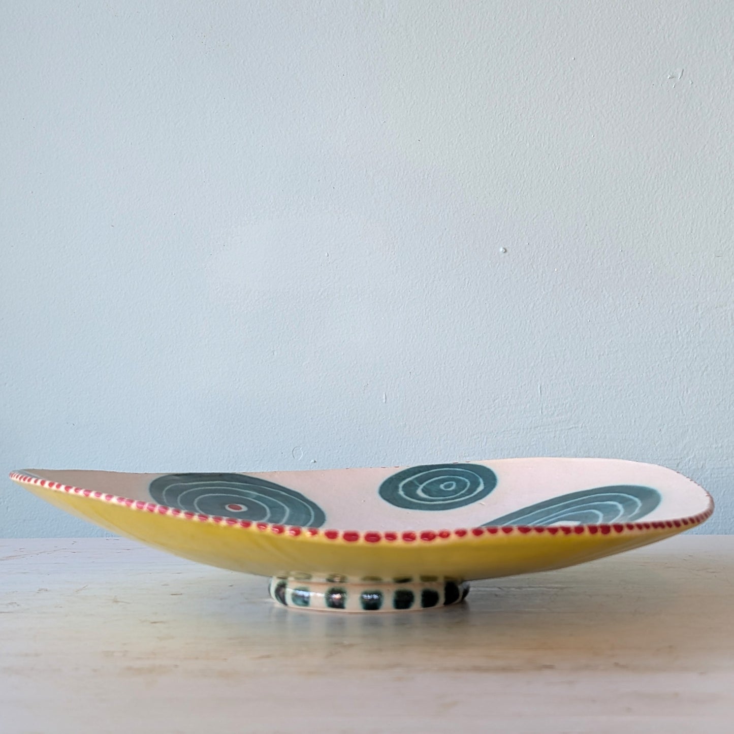A shallow ceramic serving platter with a yellow base, decorated with dark green spiral patterns and a red dotted rim, resting on a light-colored wooden surface against a pale blue wall.