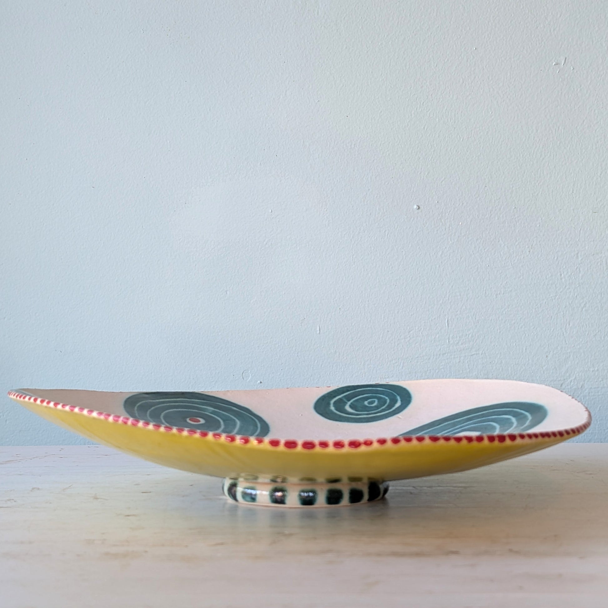 A shallow ceramic serving platter with a yellow base, decorated with dark green spiral patterns and a red dotted rim, resting on a light-colored wooden surface against a pale blue wall.