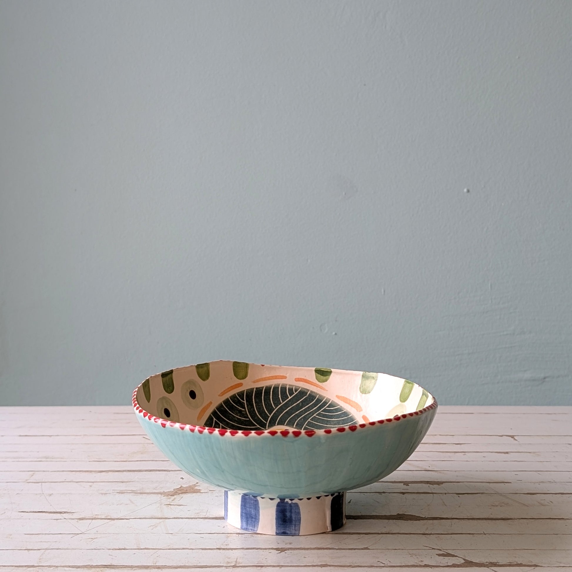 Front view of a colourful ceramic bowl with a foot ring, on a white wooden surface with a blue background.