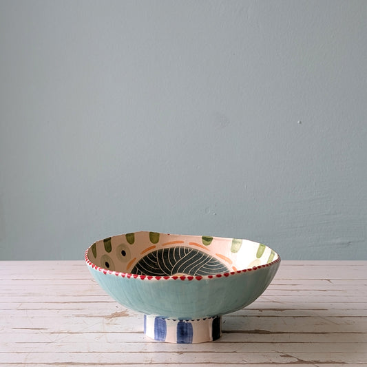 Front view of a colourful ceramic bowl with a foot ring, on a white wooden surface with a blue background.