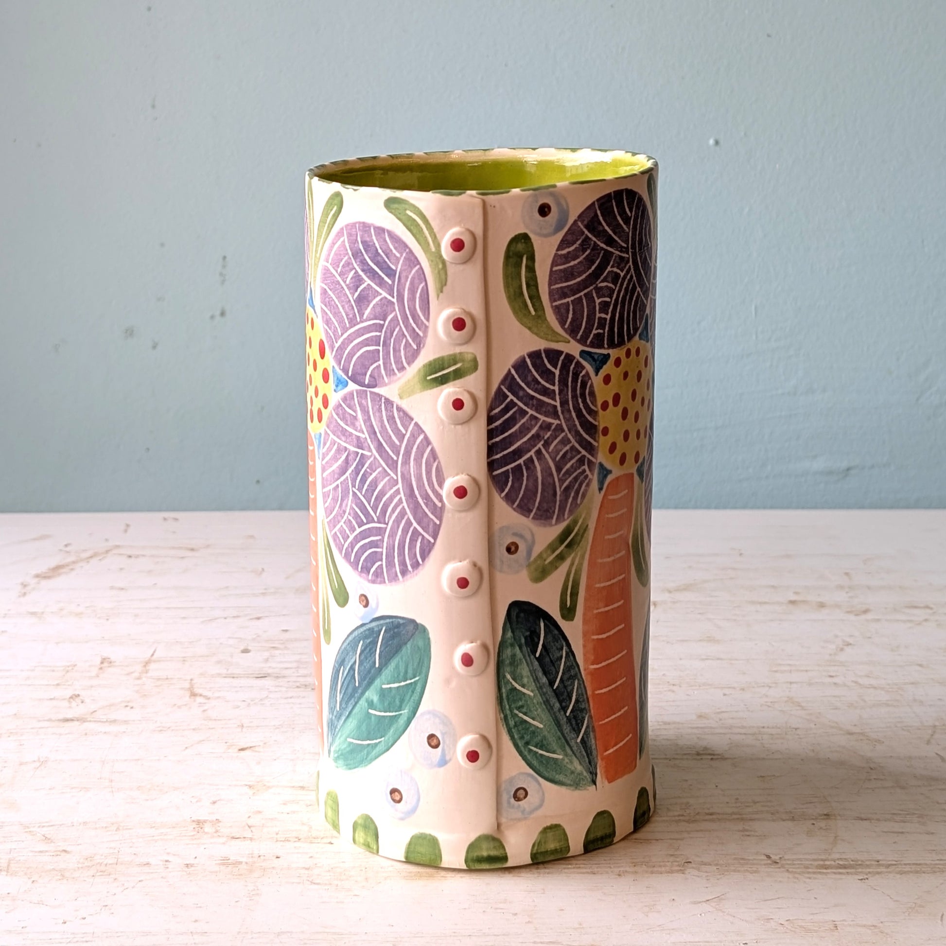 Cylindrical ceramic vase with floral patterns on a light wooden surface and blue background