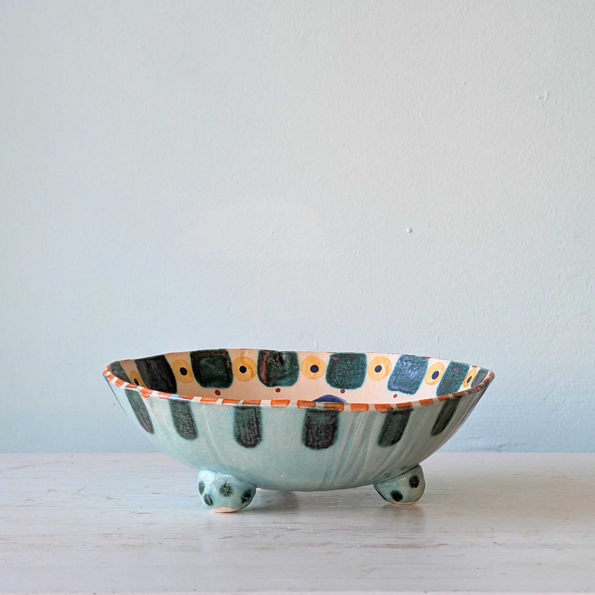 A handmade ceramic bowl with a light blue-green base, decorated with dark green and orange rectangular patterns on the interior and exterior, resting on a light wooden surface against a neutral wall.