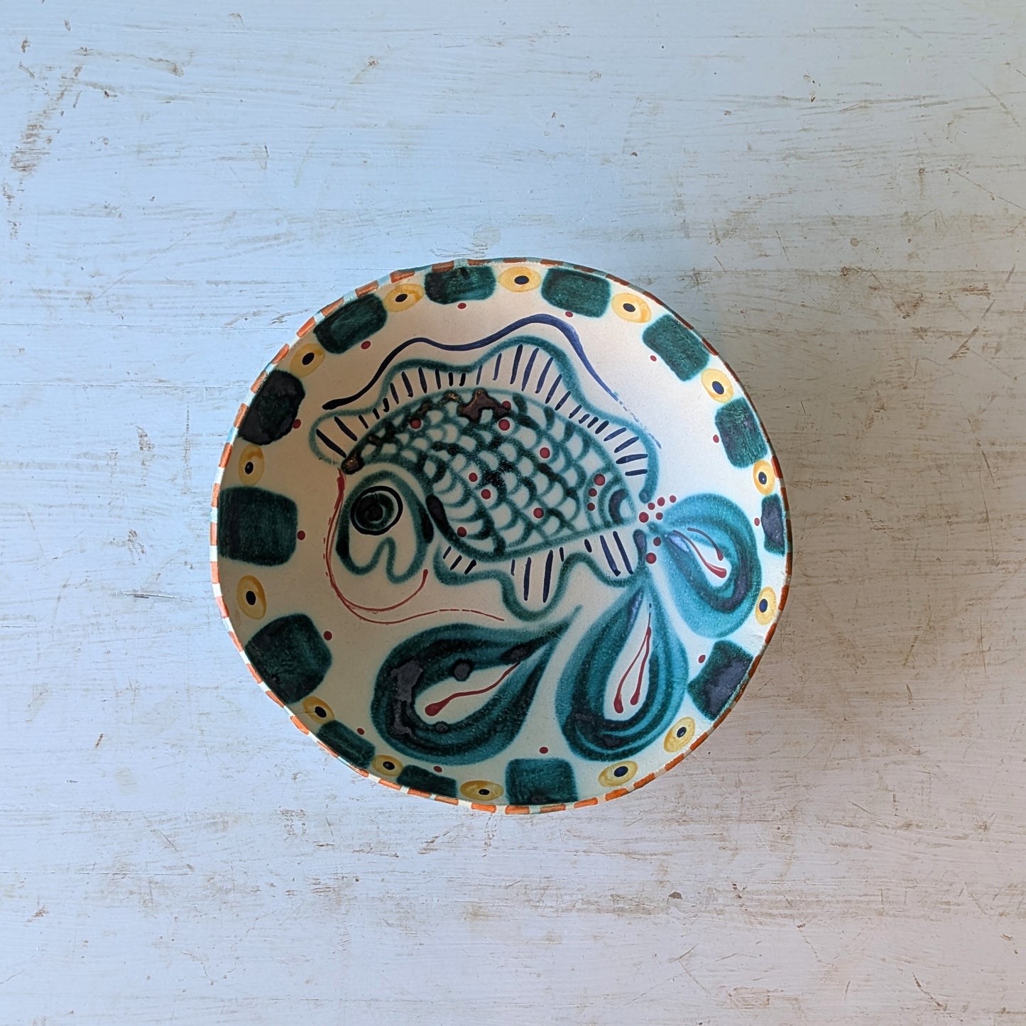 A hand-painted ceramic bowl viewed from directly above, featuring a stylized fish design in teal, green, and white, with a patterned border, set on a distressed white wooden surface.