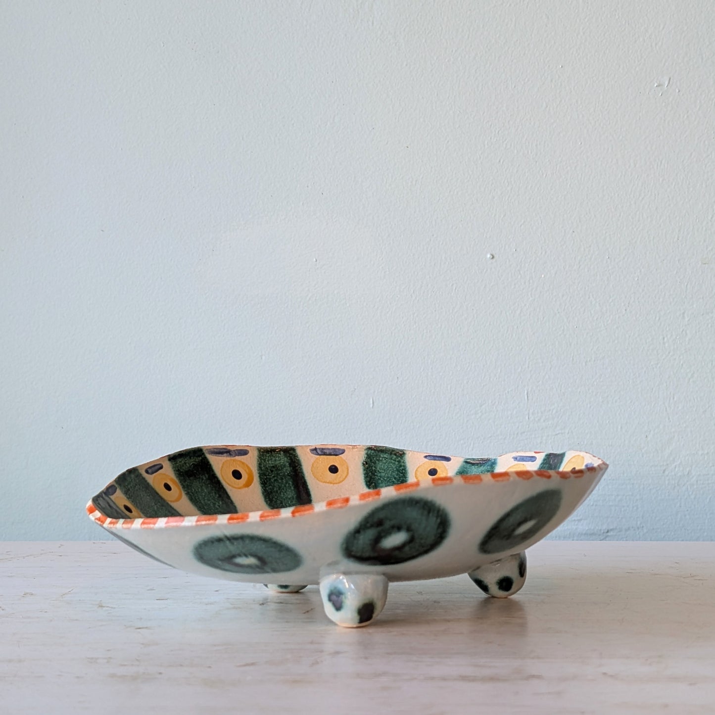 A handmade ceramic bowl with three small feet, featuring a white base decorated with green circular patterns and a striped pattern on the rim with yellow and green accents, set on a light wooden surface against a plain light blue wall.