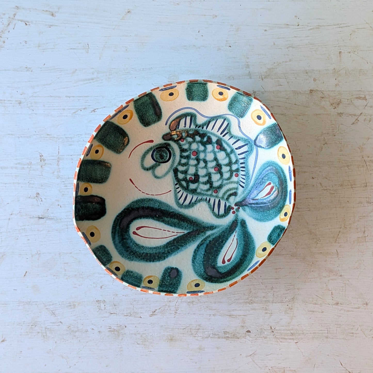 A hand-painted ceramic bowl with a stylized fish design in green, blue, and white, set on a distressed white wooden surface.