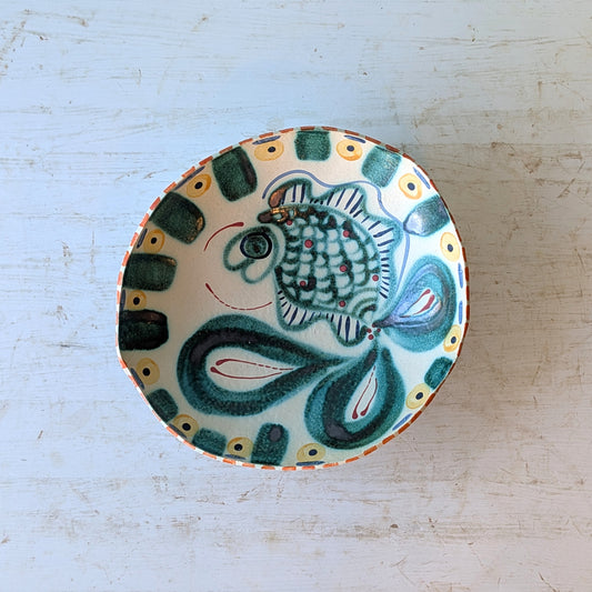 A hand-painted ceramic bowl with a stylized fish design in green, blue, and white, set on a distressed white wooden surface.