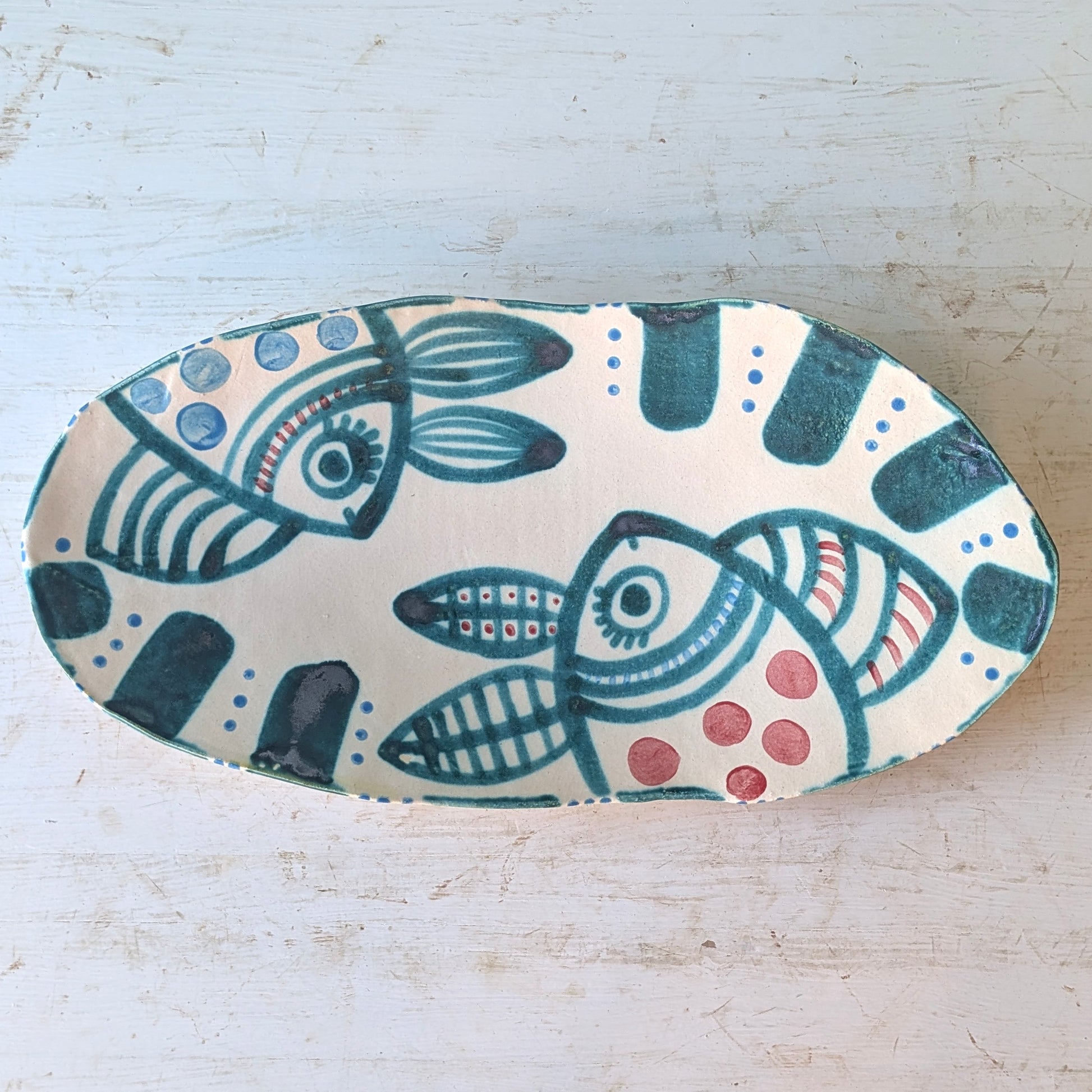 An oval ceramic serving platter with a hand-painted folk art design of stylized fish in teal, dark green, red, and blue on a cream background, set on a distressed white wooden surface.