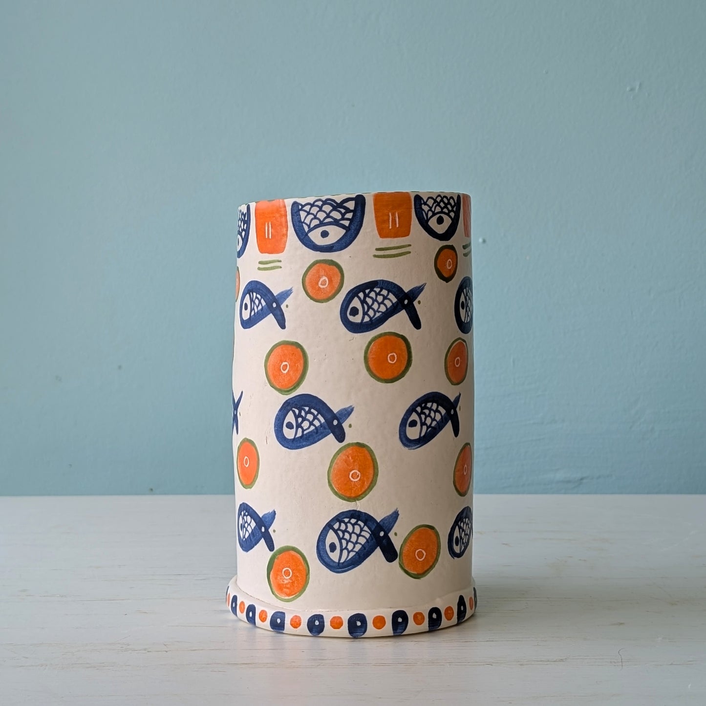 White ceramic vase with blue fish and orange circle pattern on a white table against a blue wall
